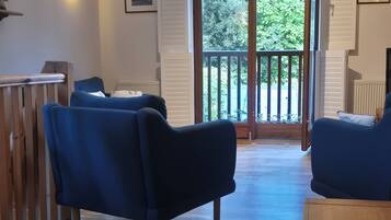 A room with a wooden floor, two blue sofas, a wooden table, and a balcony with a view of trees.