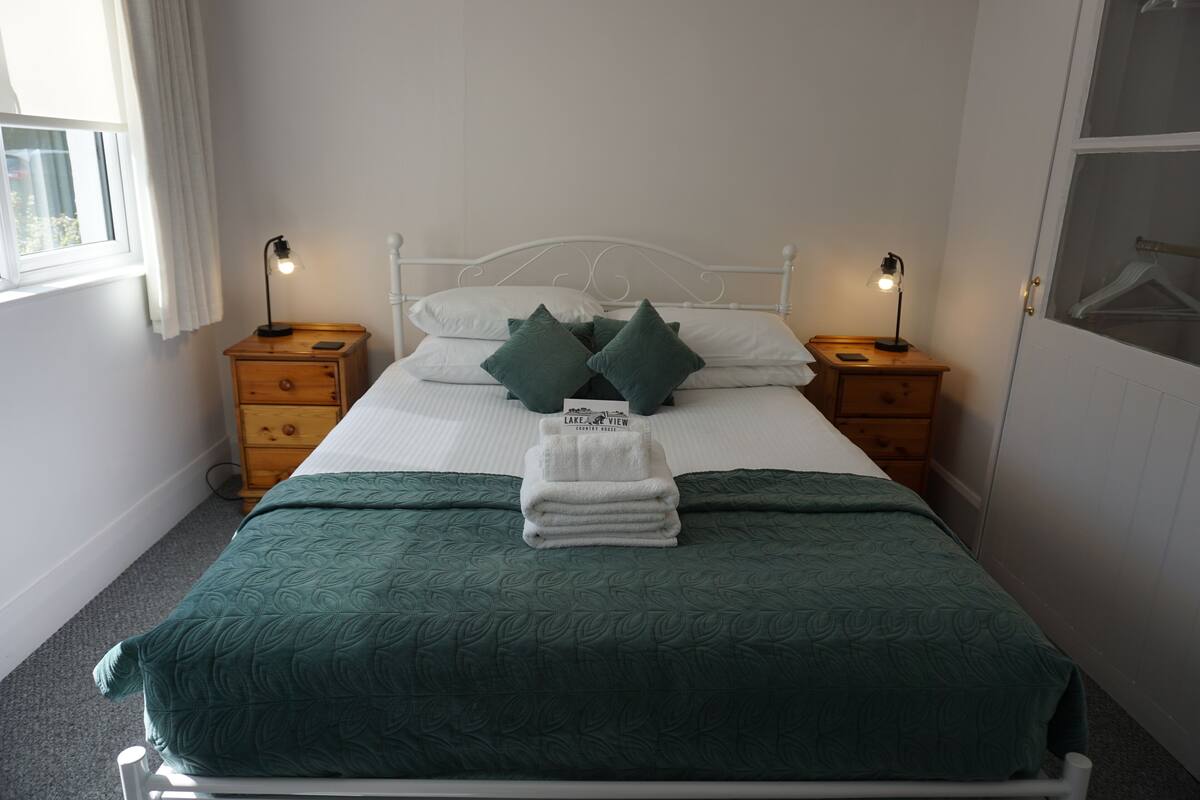 A neatly made bed with a green bedspread and pillows, two bedside tables with lamps, a wooden nightstand, and a window with curtains.