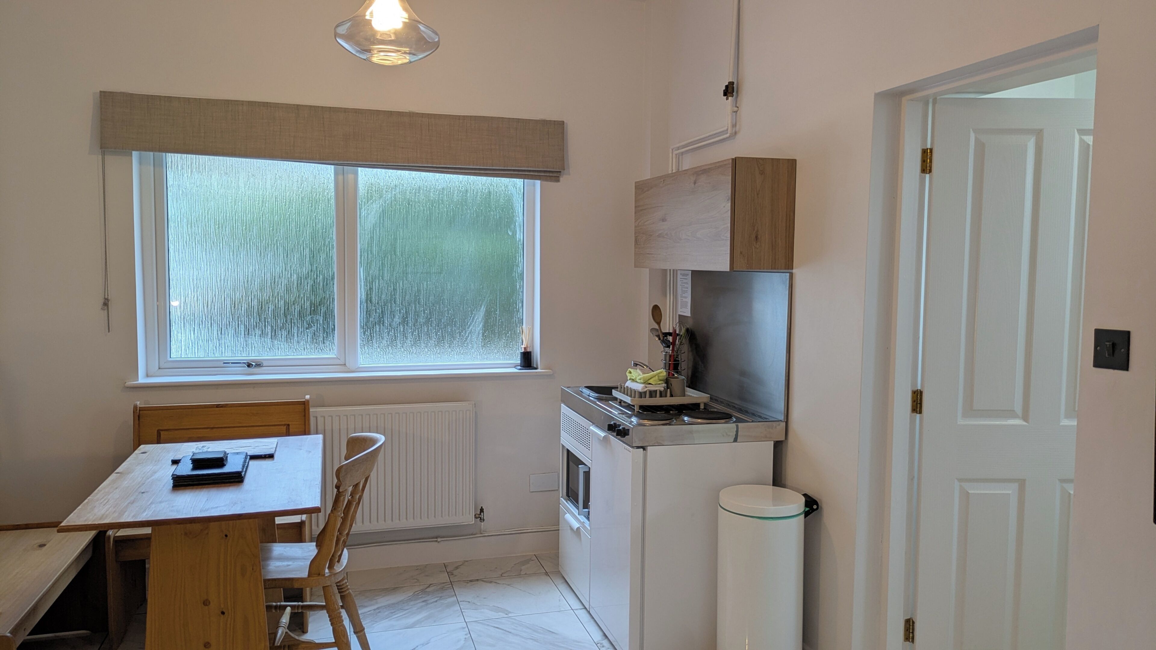 A small kitchen with a dining table, chairs, a window with blinds, a stove, and a trash bin.