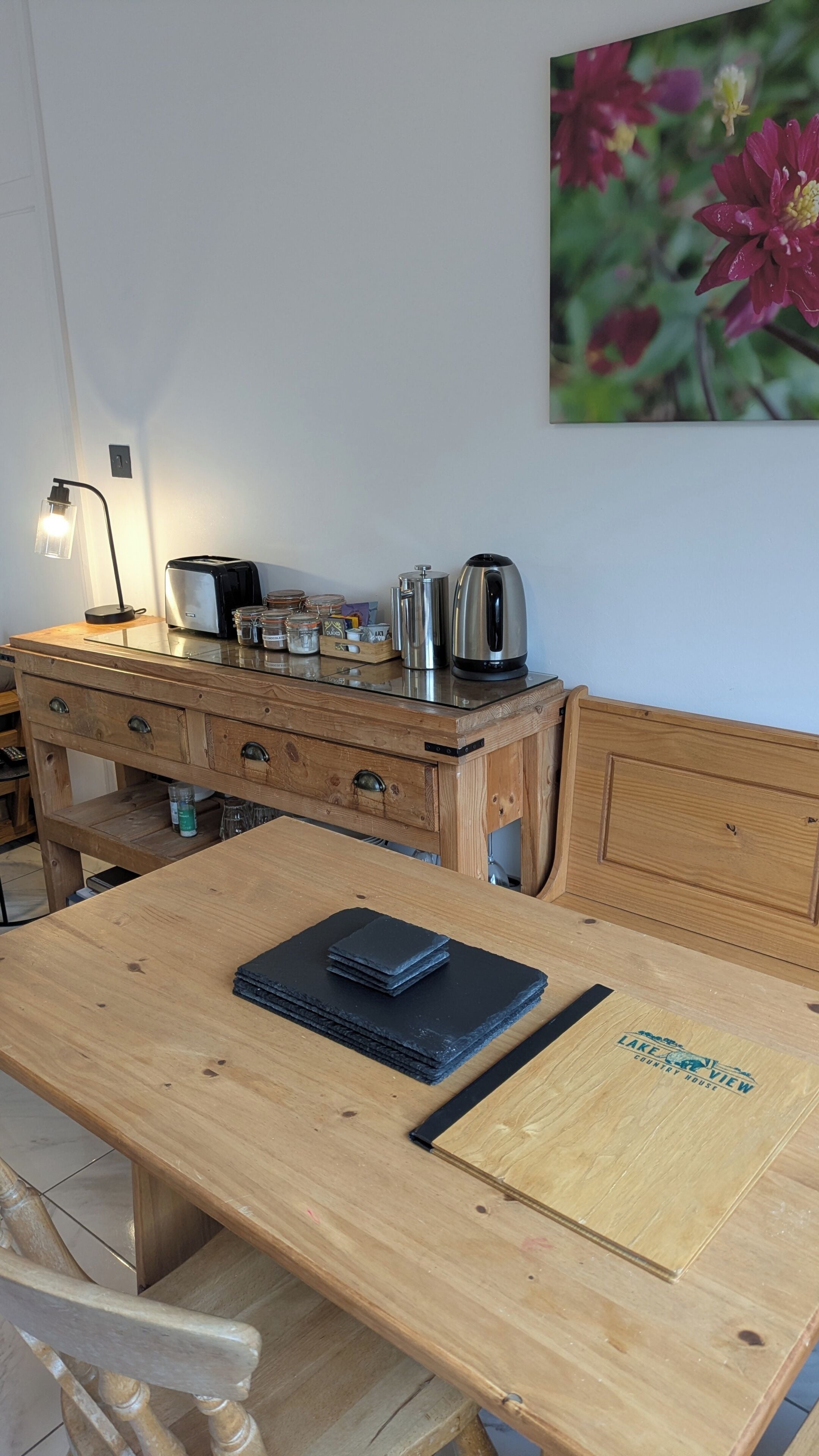 Una mesa de comedor de madera con una libreta negra, una tetera y frascos sobre un mueble de madera.