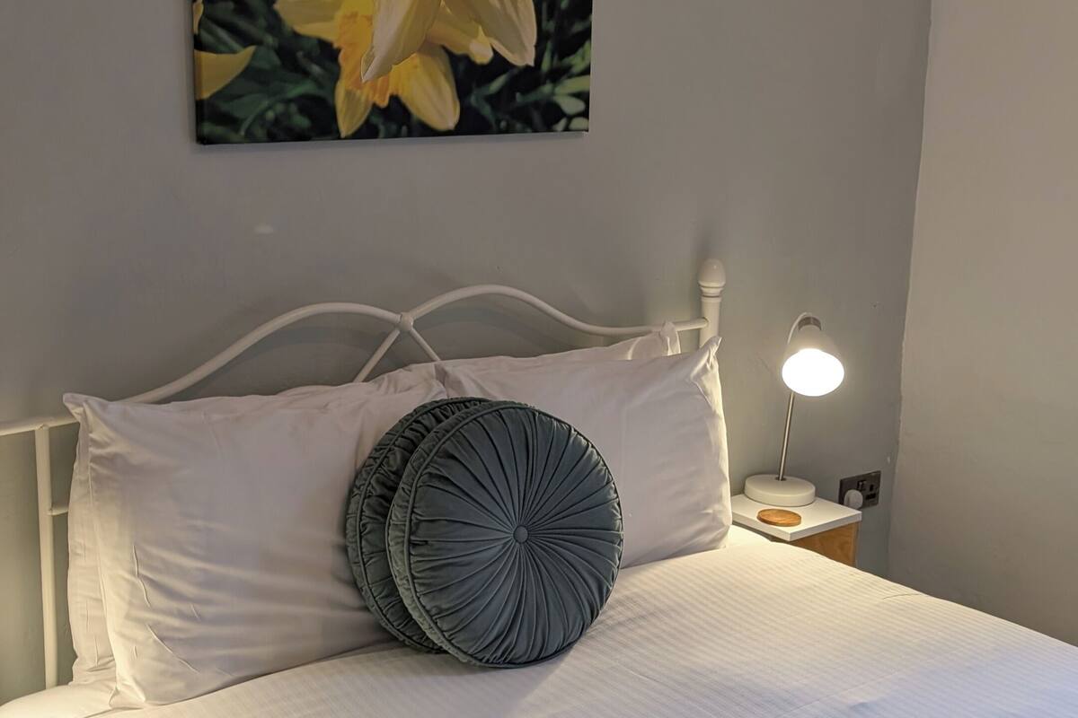 A bed with white bedding and a single gray pillow, a white bedside table with a lamp, and a painting of yellow flowers on the wall.