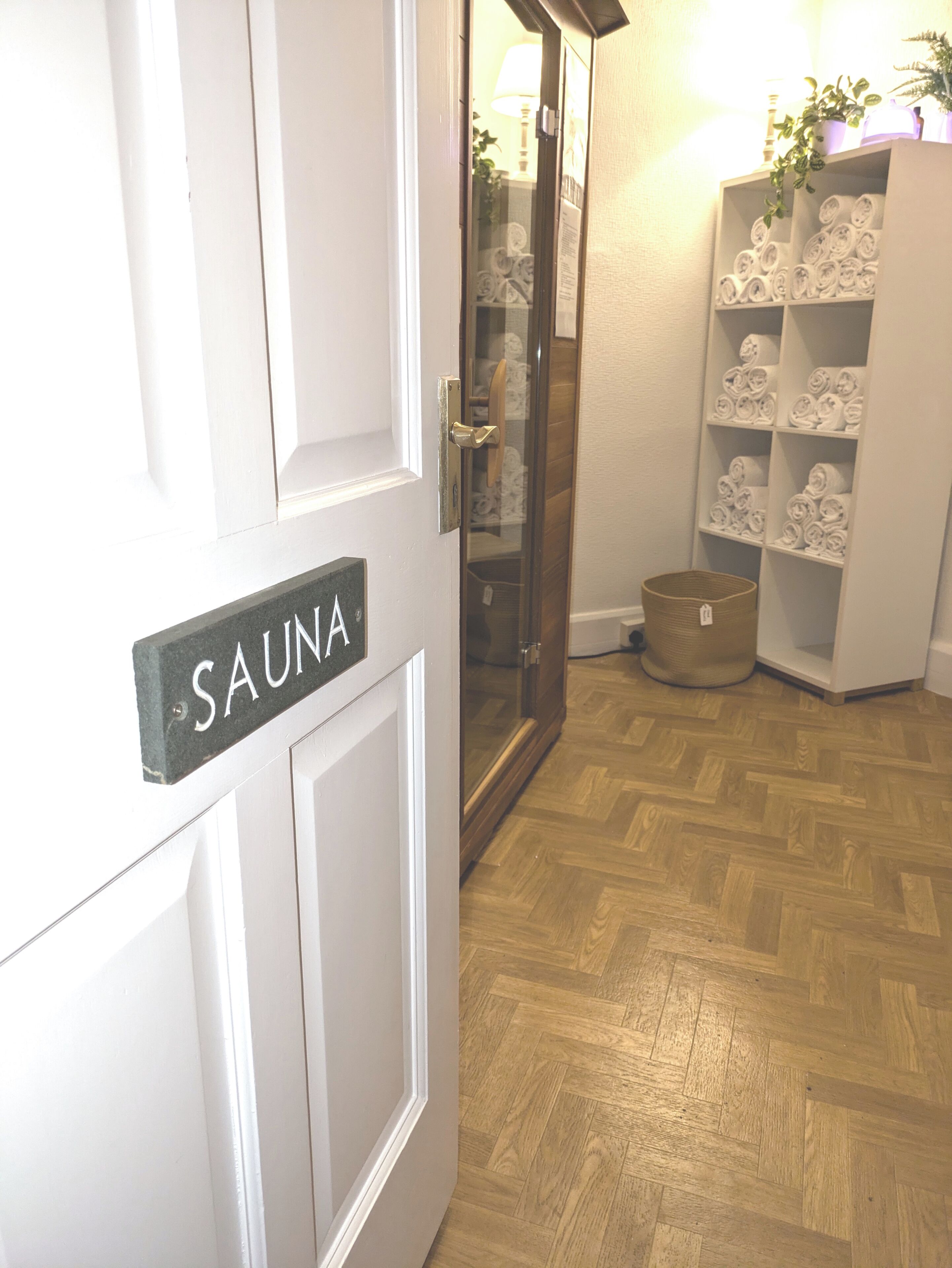 A door labeled 'SAUNA' with a wooden floor, a glass door, and a white shelving unit with towels.
