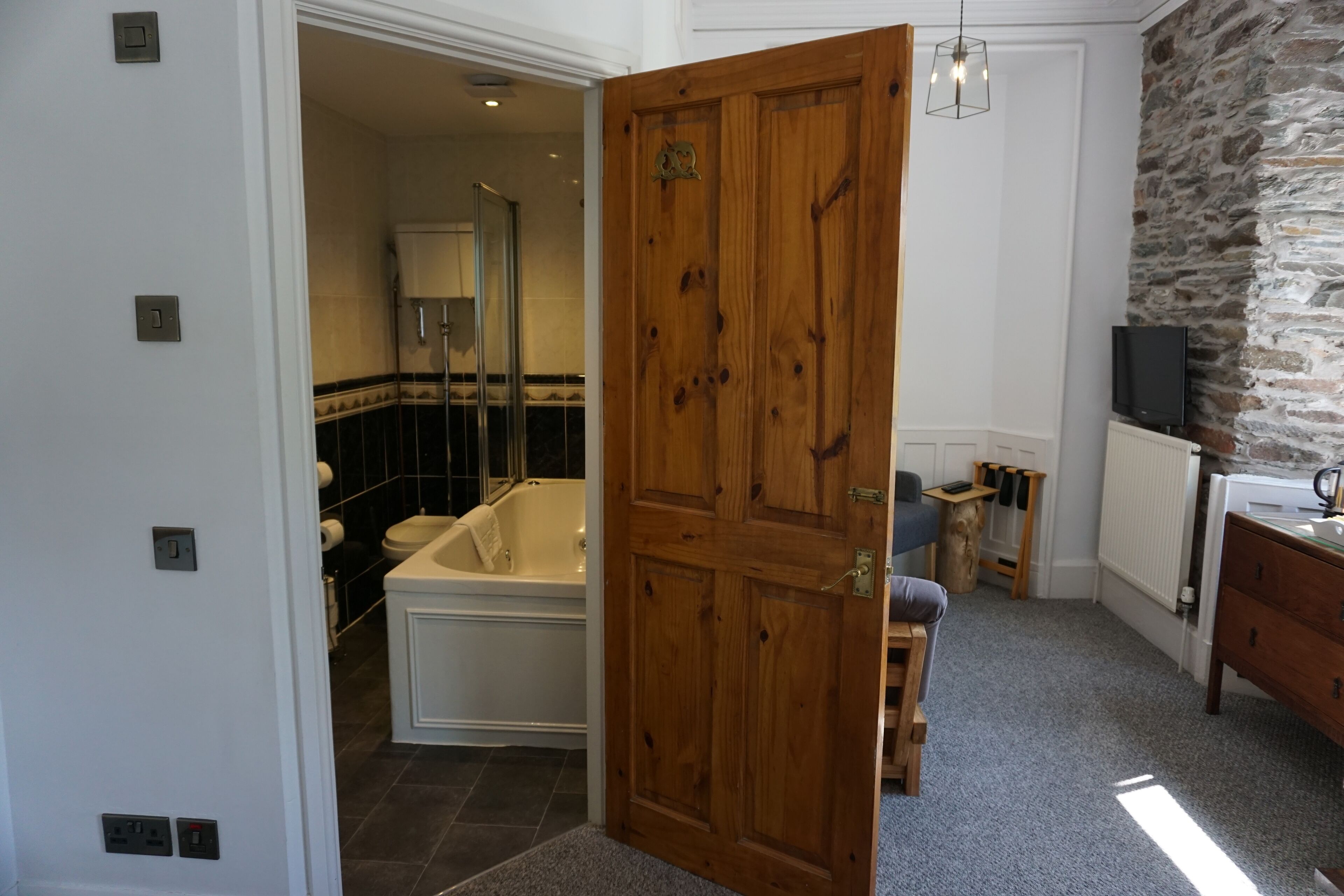 A bathroom with a bathtub, a toilet, and a shower. There is a wooden door, a stone wall, and a television on a dresser.