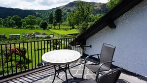 Un balcon avec une table ronde et deux chaises, donnant sur un paysage verdoyant, avec des arbres et une montagne en arrière-plan.