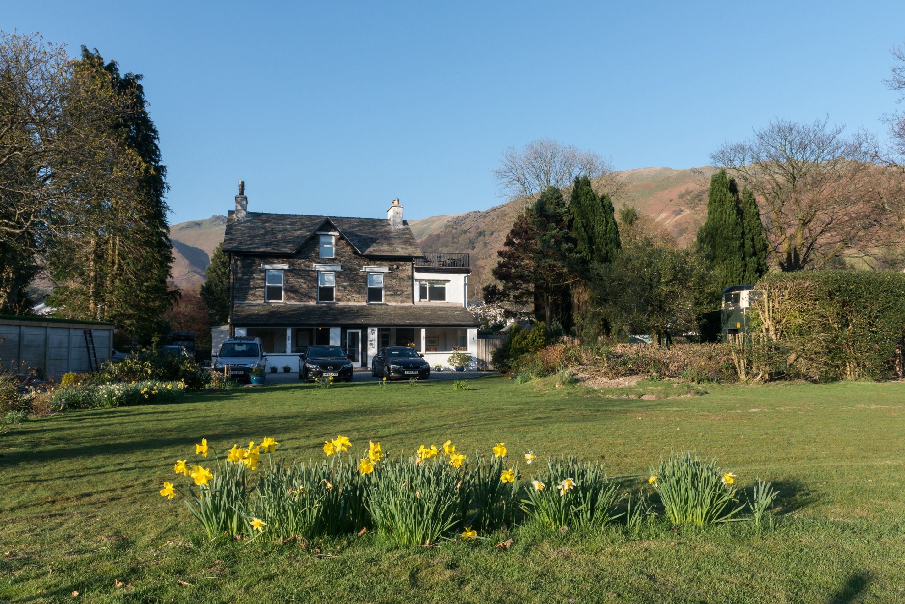A two-story stone house with a front lawn, parked cars, and a mountainous backdrop.