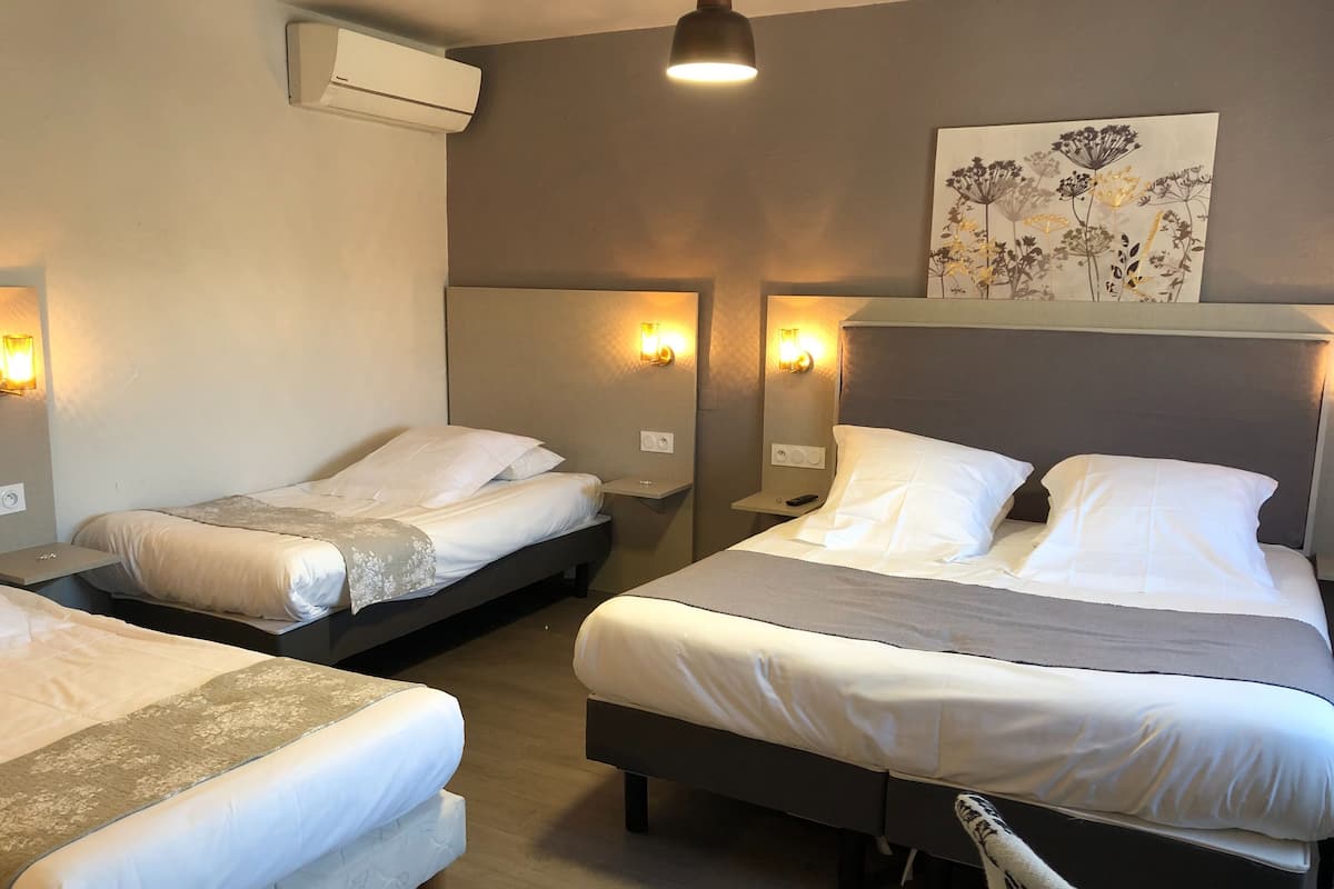 Ein Hotelzimmer mit drei Betten, jedes mit weißer Bettwäsche und einer grauen Tagesdecke. An den Wänden sind Leuchten angebracht und ein Gemälde hängt über den Betten.