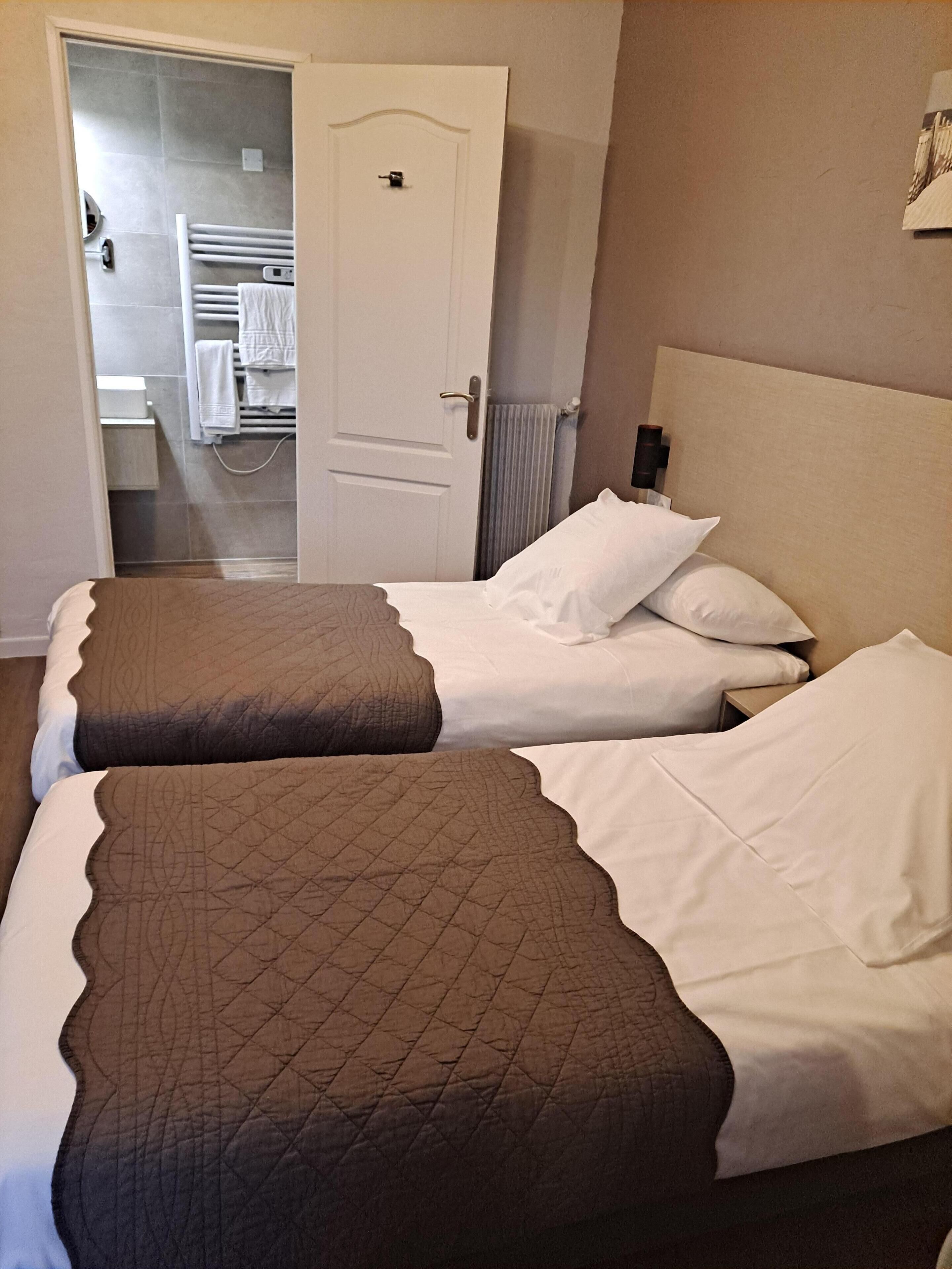 A hotel room with two beds, each with a brown quilted bedspread and white linens. A white door leads to a bathroom with a towel rack.