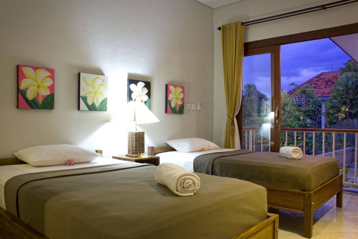 Quarto de hotel com duas camas, uma mesinha de cabeceira, luminária e uma obra de arte na parede.
