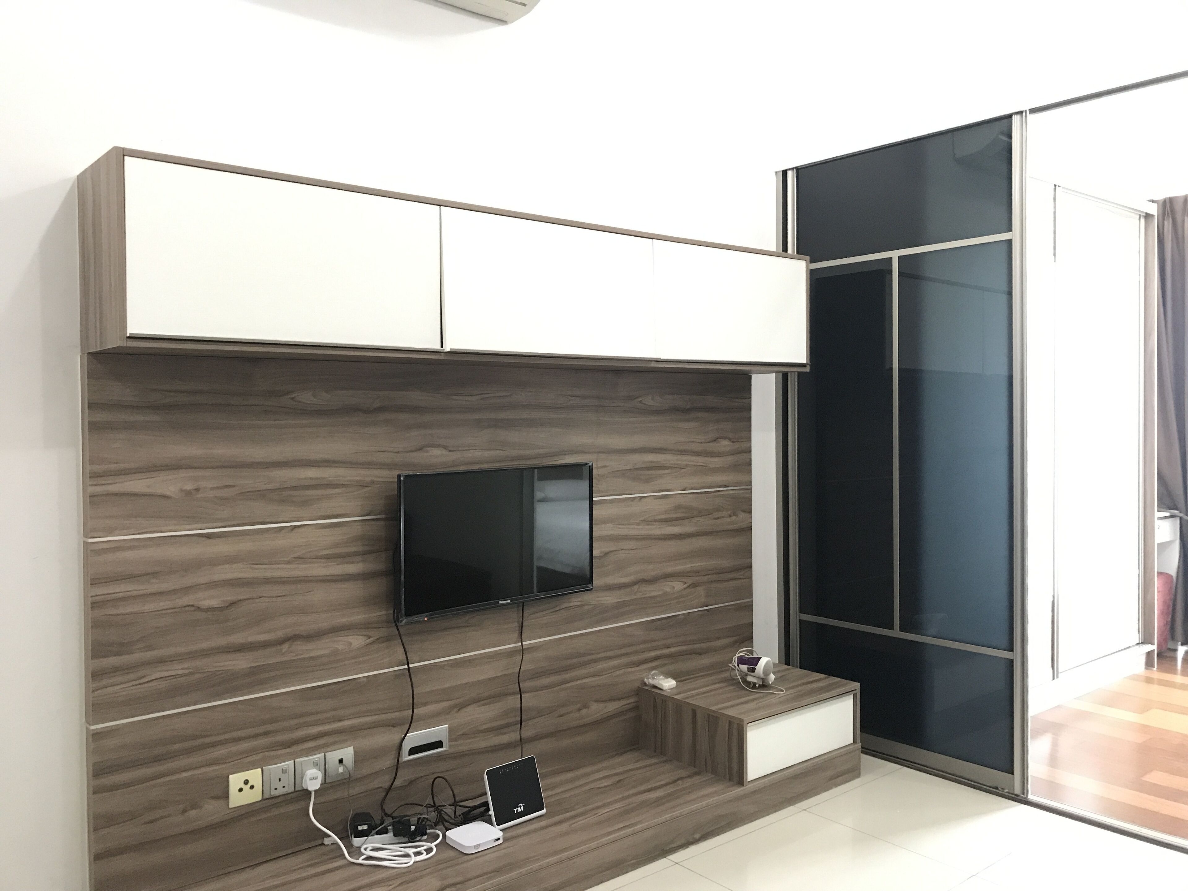 Una habitación moderna con televisión de pantalla plana en una pared de madera, una mesita con un altavoz y una puerta corredera de cristal que da a un balcón.