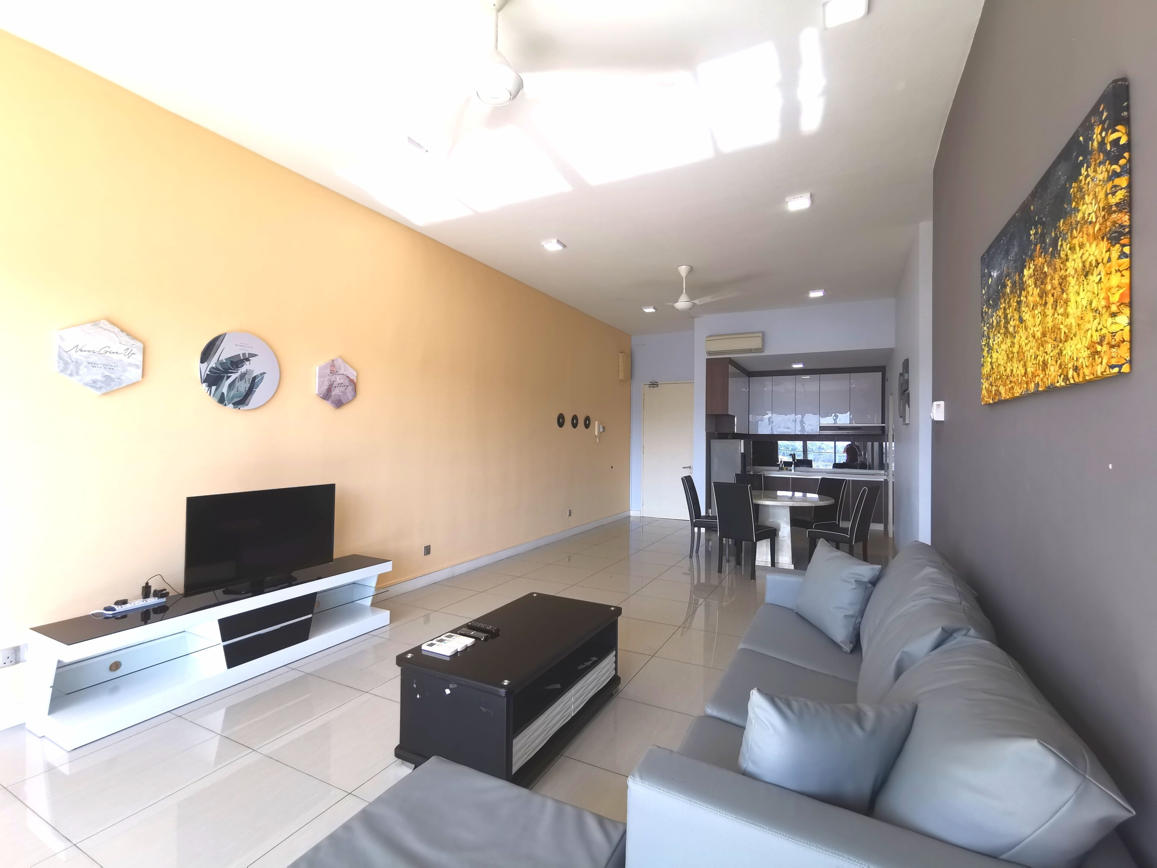 A modern living room with a flat-screen TV, a sofa, and a coffee table. There is a kitchen area in the background with a dining table and chairs.