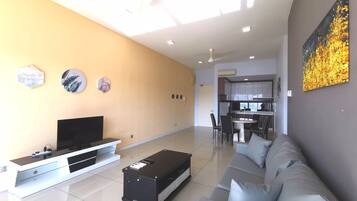 A modern living room with a flat-screen TV, a sofa, and a coffee table. There is a kitchen area in the background with a dining table and chairs.