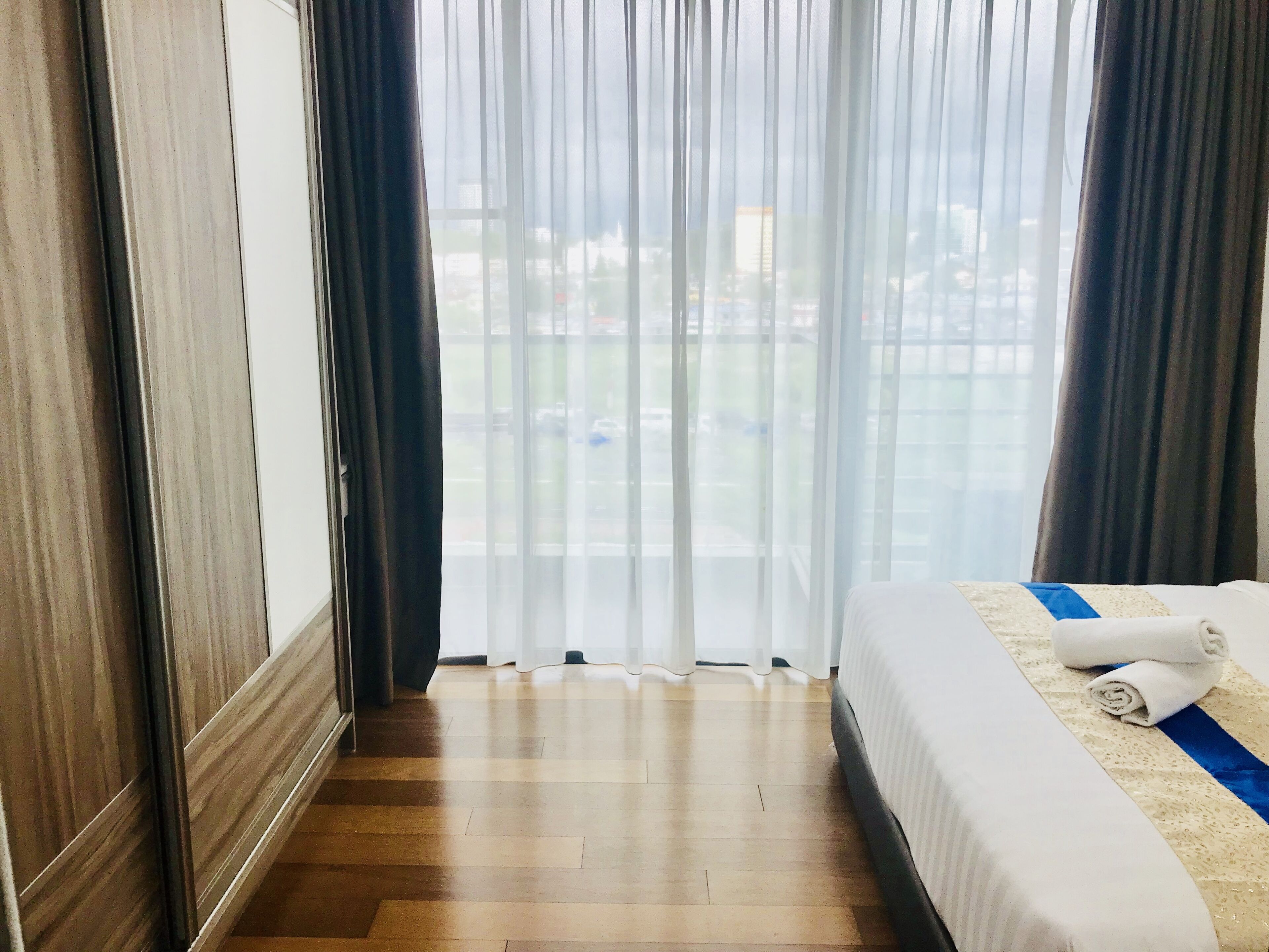 Una habitació d'hotel amb una finestra gran, terres de fusta, un llit ben fet amb una tovallola arrotllada i vista a un paisatge urbà.