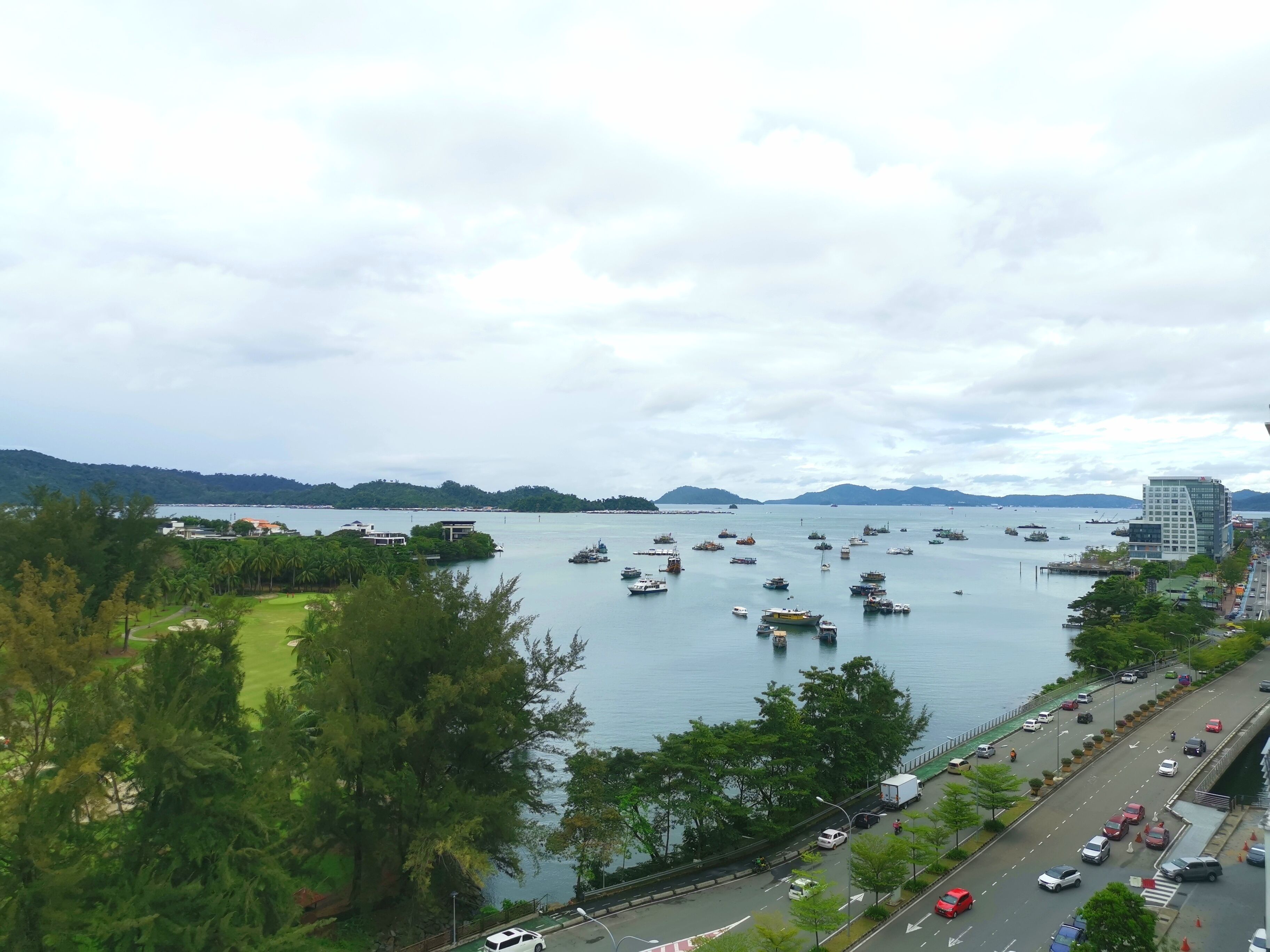 Un paysage urbain côtier avec un port de plaisance rempli de bateaux, une route où circulent des véhicules et des bâtiments le long du front de mer.