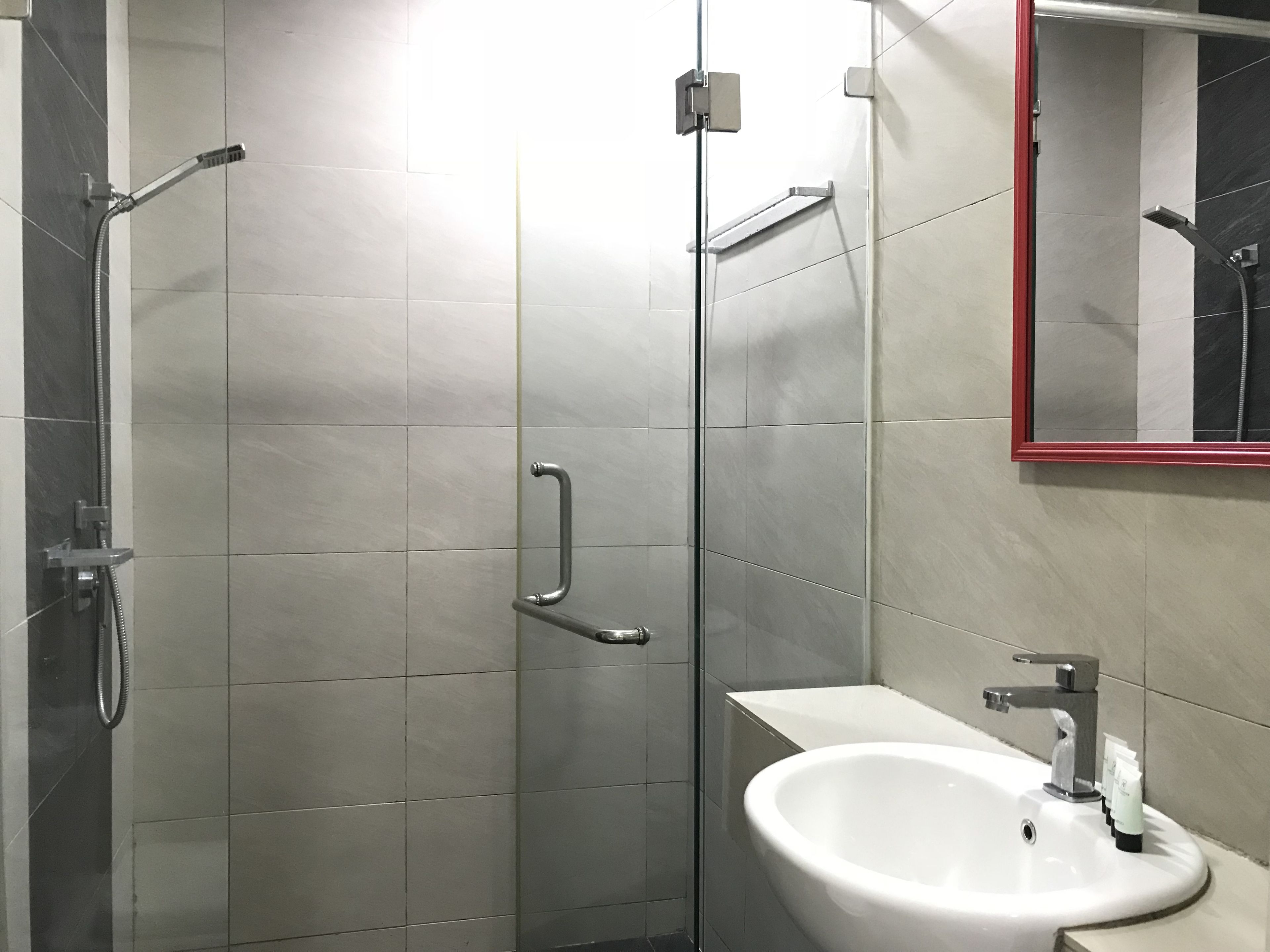 Un baño moderno con ducha envuelta en vidrio, un lavabo blanco y un espejo con marco rojo.