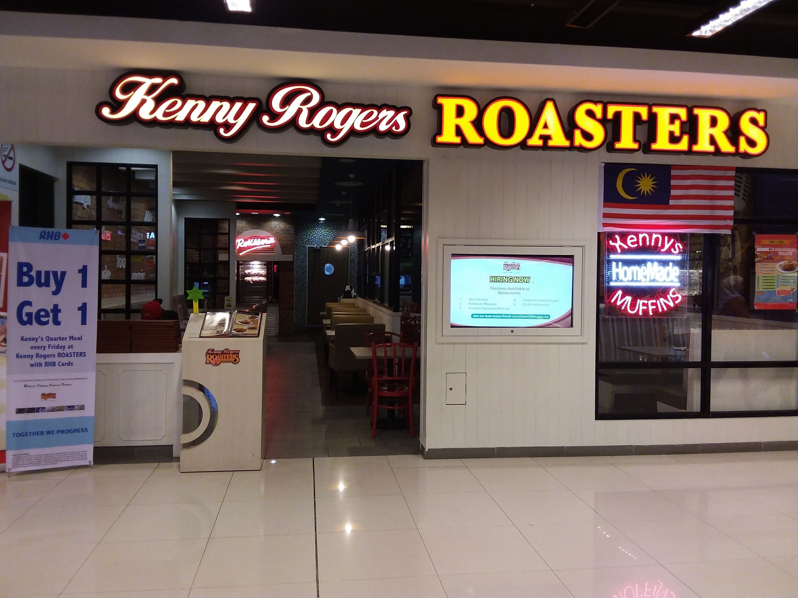Un restaurant Kenny Rogers Roasters avec une enseigne au néon, une affiche d’offre d’emploi et une promotion du type « achetez-en un, obtenez-en un gratuitement ».