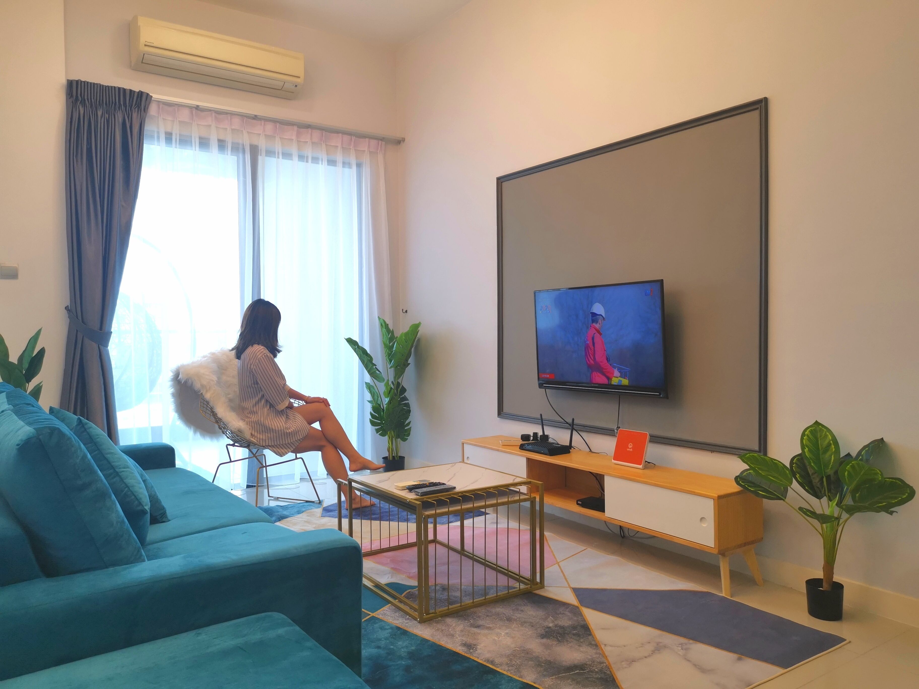 A modern living room with a large flat-screen TV mounted on the wall, a blue sofa, a small coffee table, and two potted plants.