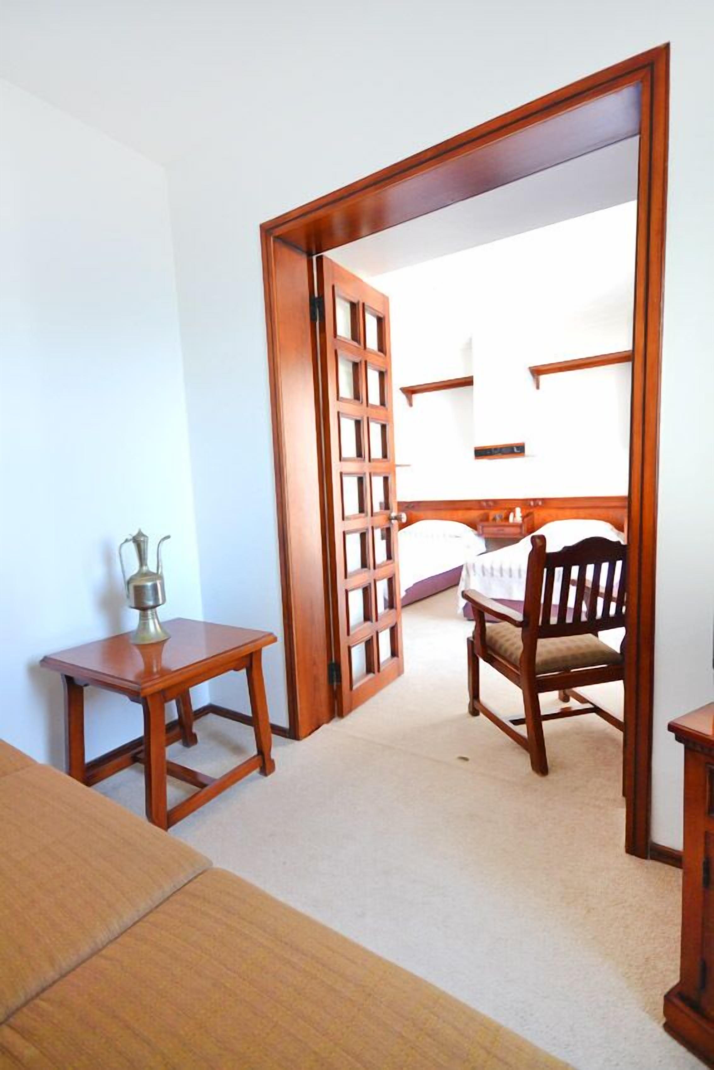 Una habitación con puerta de madera, una mesita con jarra metálica, una silla de madera y una cama con cabecera blanca.