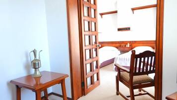 Una habitación con puerta de madera, una mesita con jarra metálica, una silla de madera y una cama con cabecera blanca.
