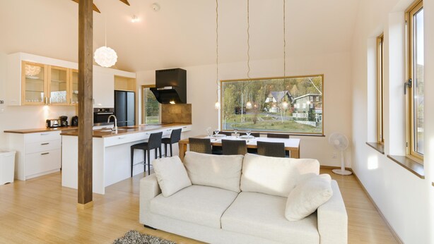 A modern kitchen with a central island, a dining area with a white sofa, and large windows offering a view of a residential area. - Hana & Jo (Kutchan)