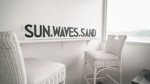 Una habitación con dos sillas blancas de mimbre, un letrero en la pared que dice "SUN.WAVES.SAND" y vista al mar a través de una ventana.