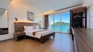 Una habitación de hotel con cama, mesita de noche, escritorio, silla, ventana grande con vista al mar y armario.