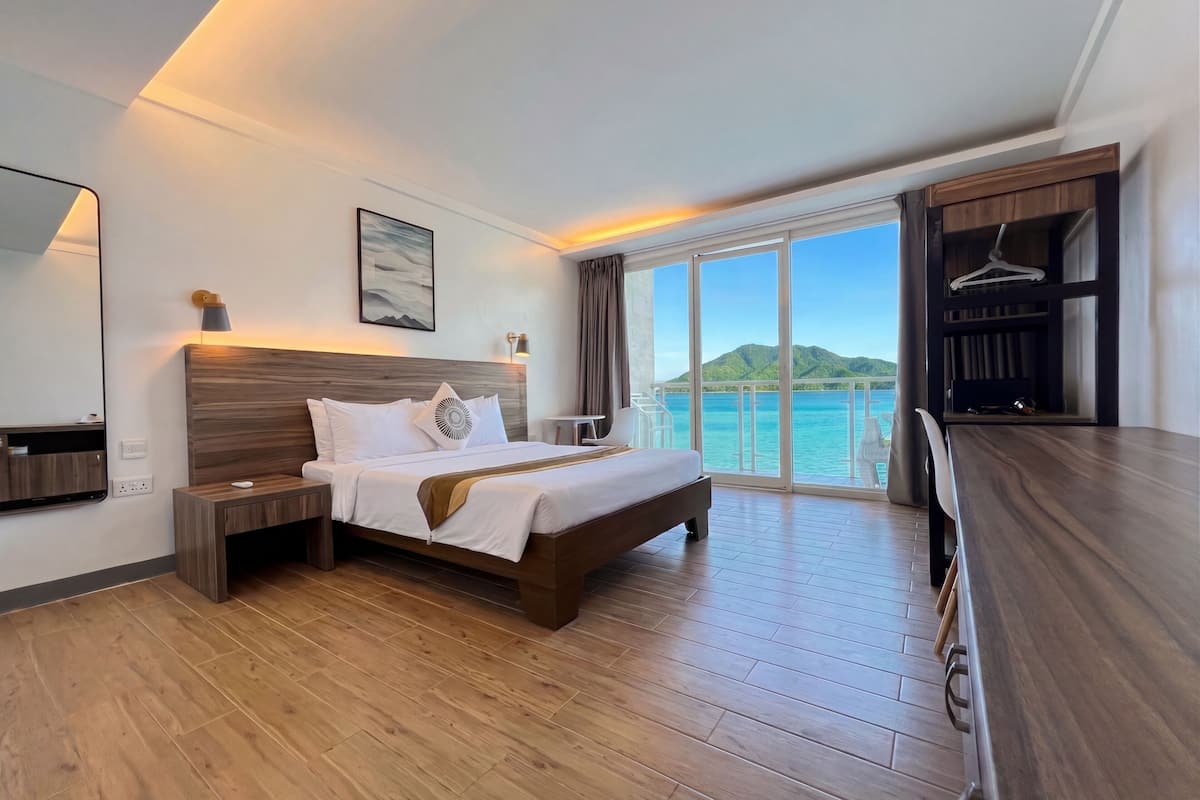 A hotel room with a bed, bedside table, a desk, a chair, a large window with a view of the sea, and a wardrobe.