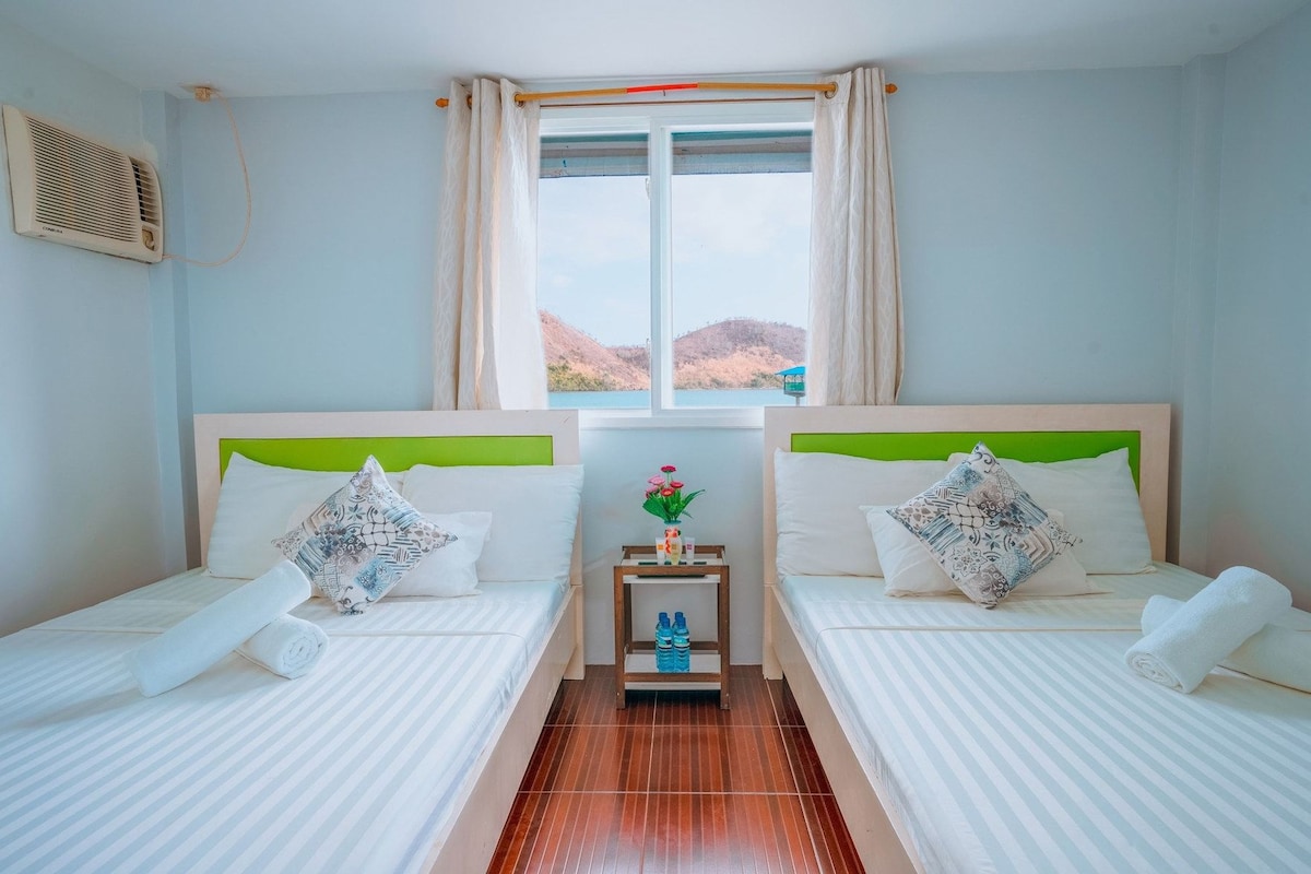 Habitación de hotel con dos camas, una ventana con cortinas, una mesita con un jarrón de flores y un equipo de aire acondicionado instalado en la pared.