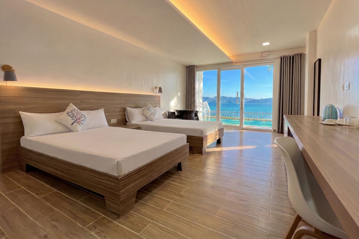 A hotel room with two beds, a wooden floor, a desk, and a view of the sea.