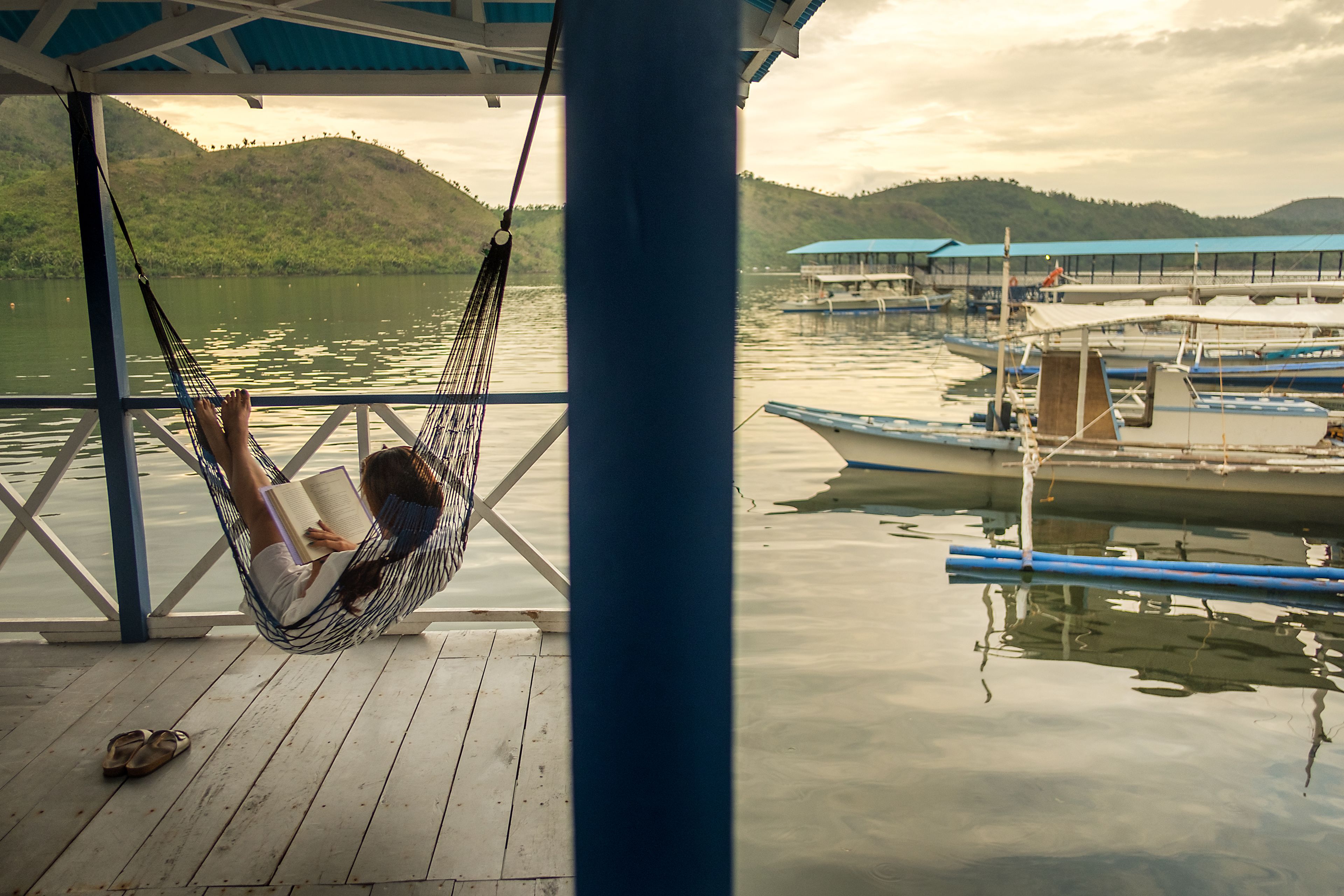 A hammock on a wooden deck overlooking calm water with boats and hills in the background.