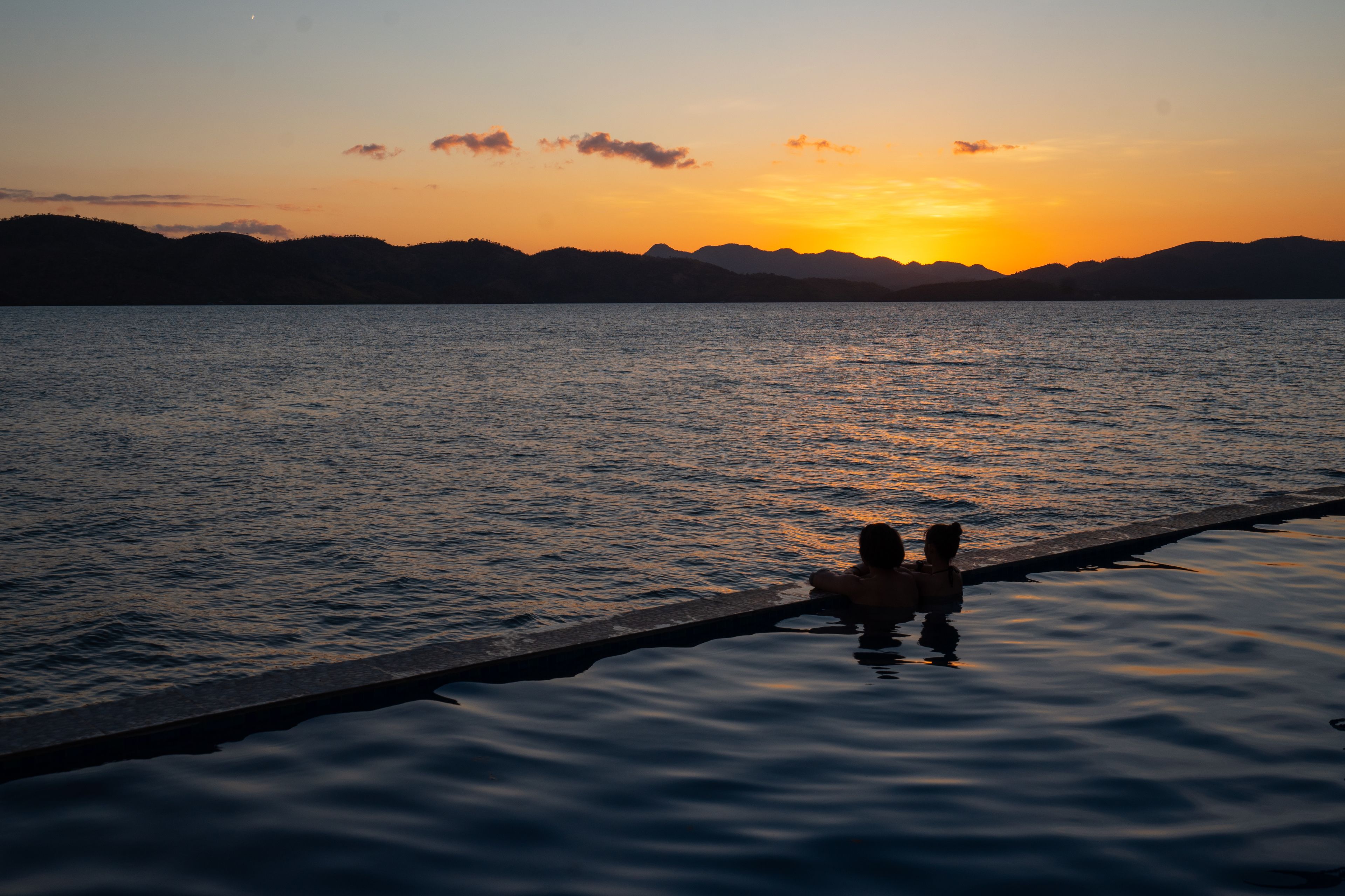 Ein Sonnenuntergang über ruhigem Wasser, im Vordergrund eine Silhouette einer Person und eines Hundes auf einem Steg.