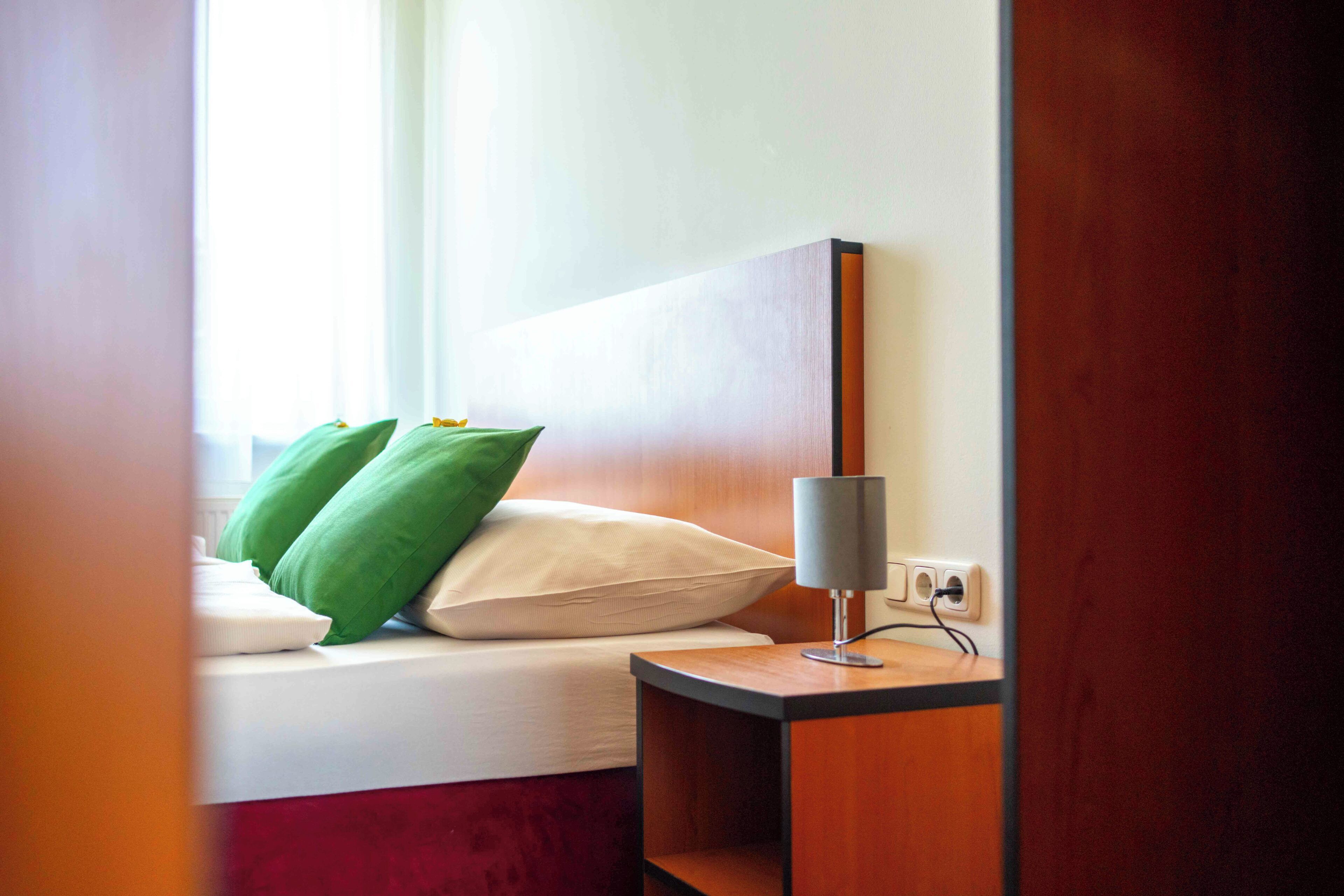 A hotel room with a bed, green and white pillows, a bedside table with a lamp, and a wooden headboard.