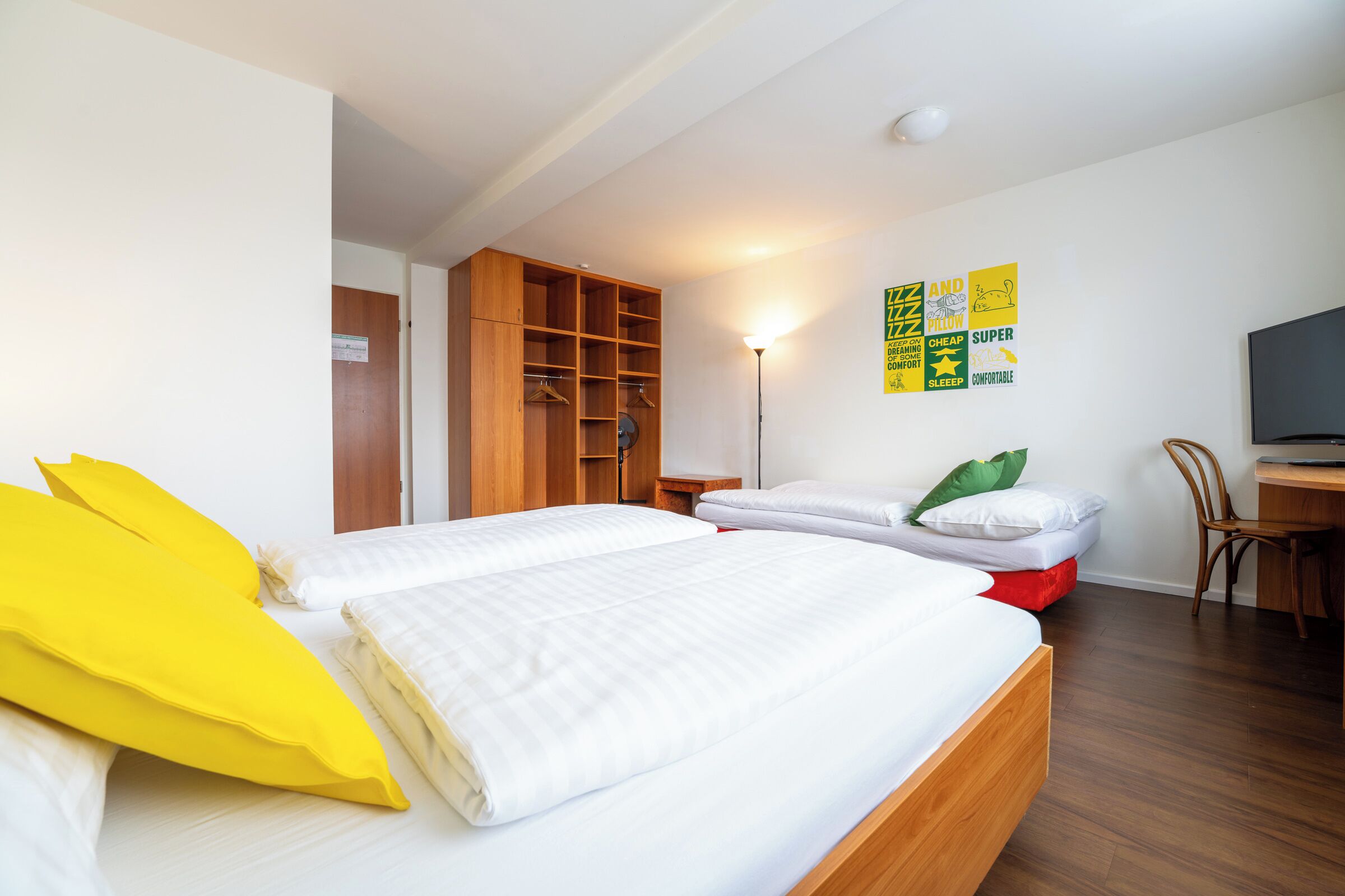 A bedroom with two beds, a wooden wardrobe, a desk with a chair, and a television.