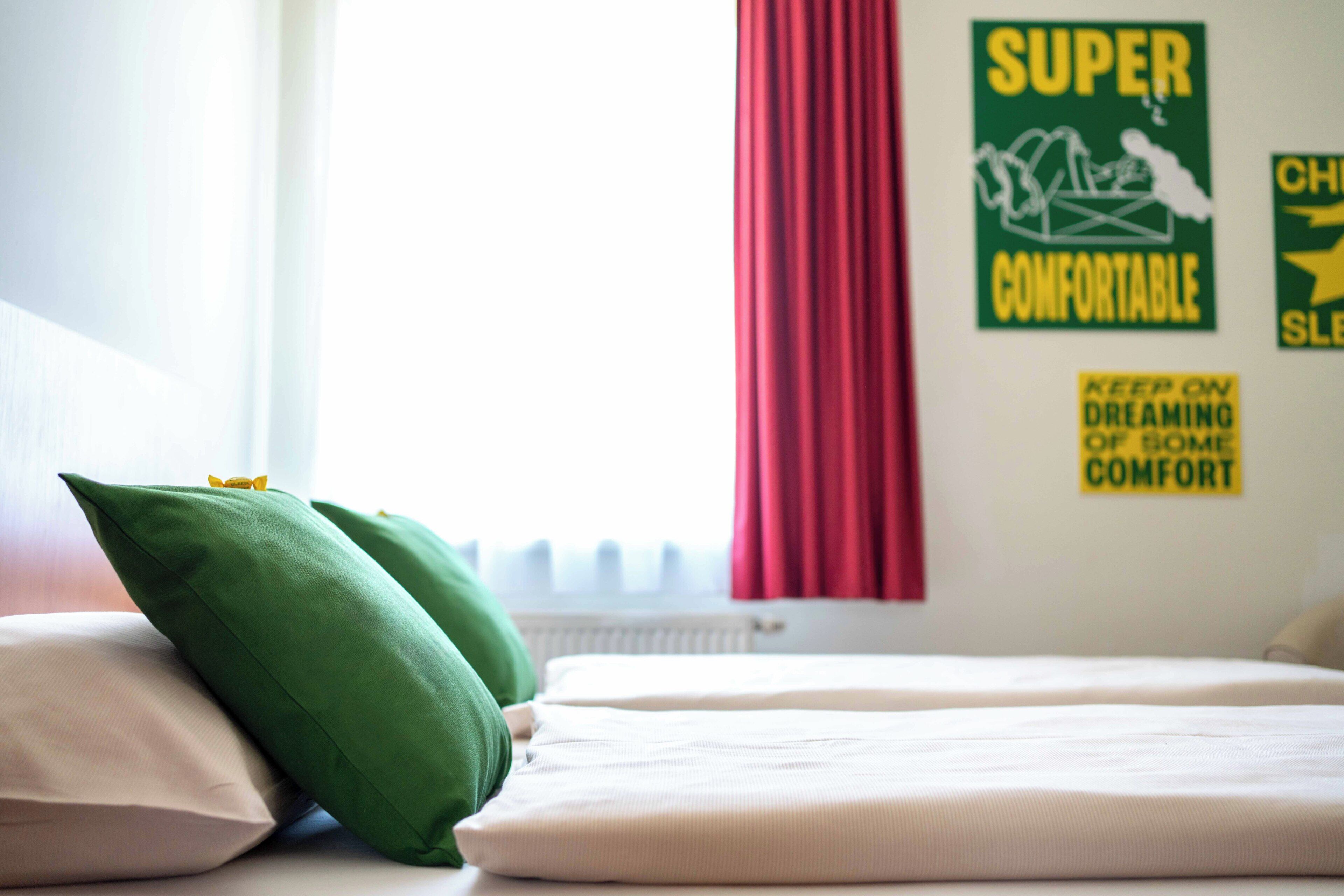 Ein ordentlich gemachtes Bett mit Kissen, ein Fenster mit Vorhängen und eine Wand mit dem Schriftzug 'SUPER COMFORTABLE'.