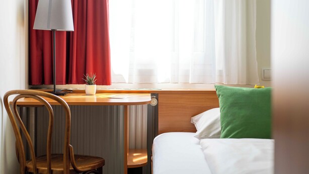 Une chambre d’hôtel avec un lit, un bureau avec une lampe, une chaise et une fenêtre avec des rideaux.