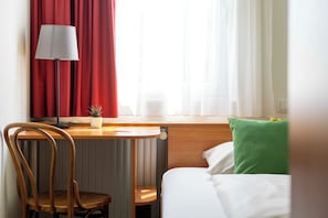 Une chambre d’hôtel avec un lit, un bureau avec une lampe, une chaise et une fenêtre avec des rideaux.
