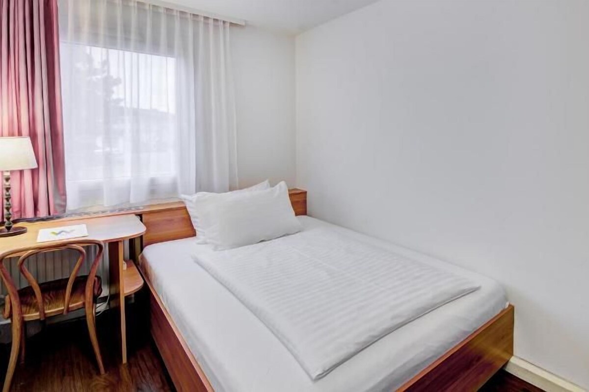 Quarto de hotel com uma cama de solteiro, uma mesa com cadeira e uma janela com cortinas.