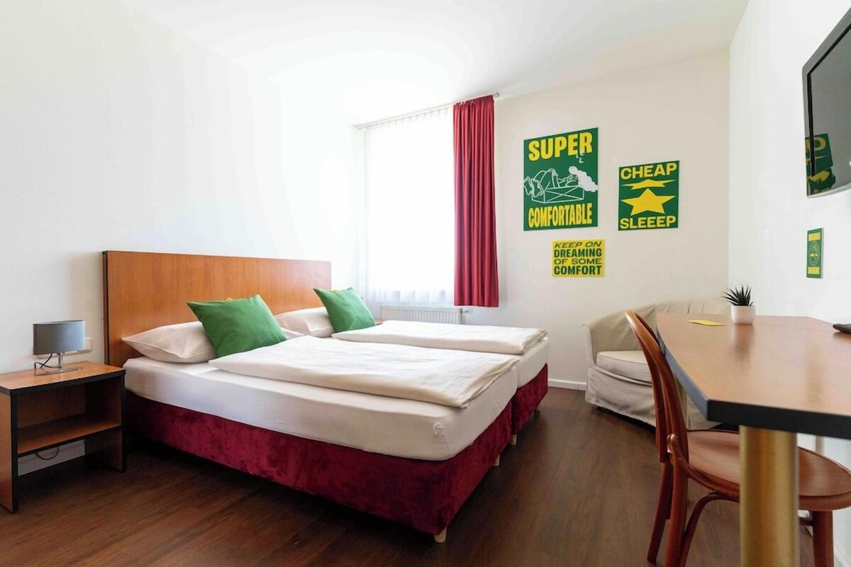 Quarto de hotel com cama de casal, pequena escrivaninha, cadeira e janela com cortinas.