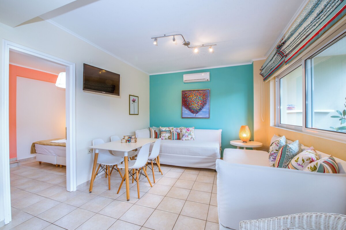 A modern living room with a white sofa, a dining table with chairs, a TV mounted on the wall, and a colorful artwork above the sofa.