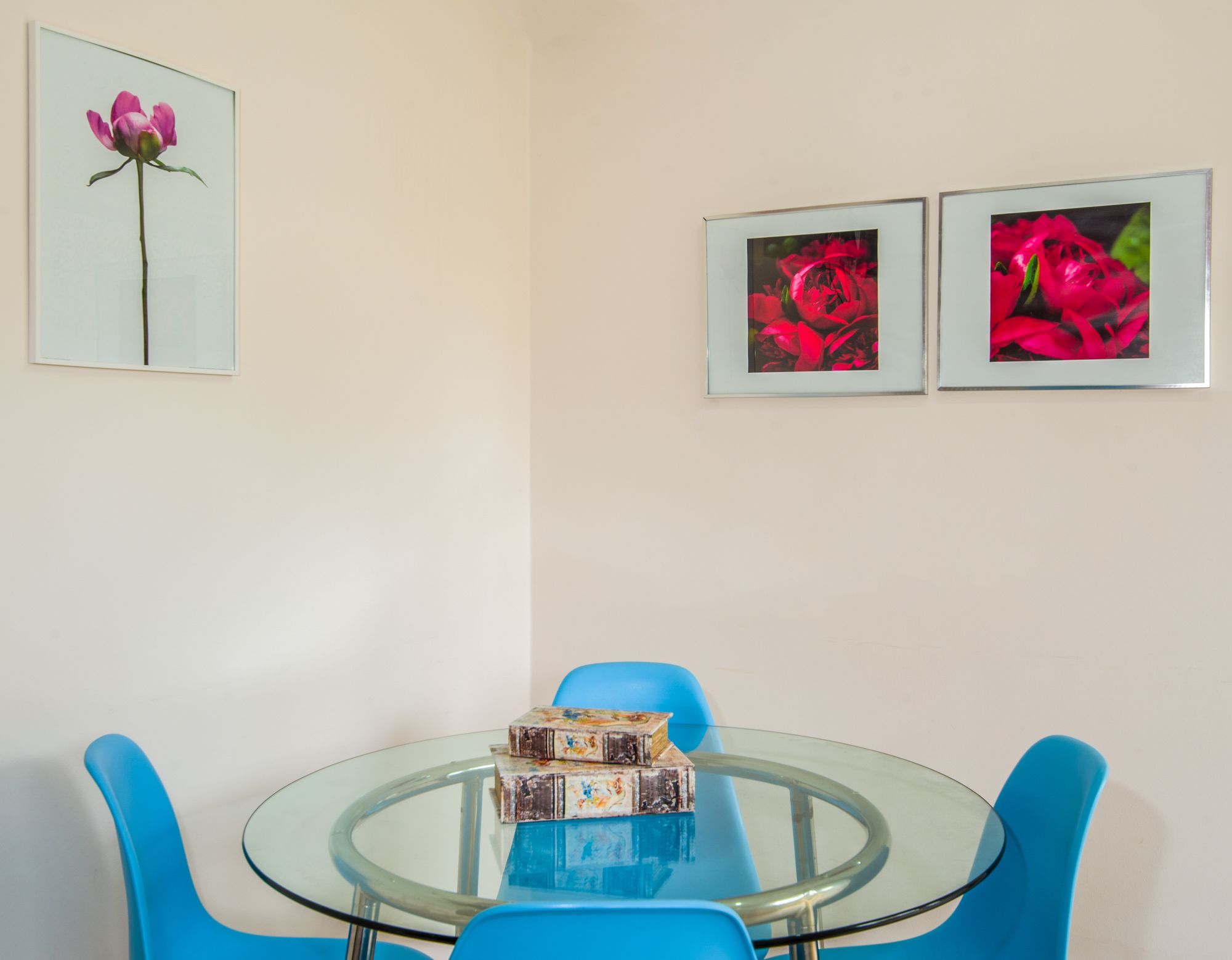 Ein Essbereich mit einem Glastisch, umgeben von blauen Stühlen, einer kleinen Auswahl dekorativer Schachteln und drei gerahmten Blumenbildern an der Wand.