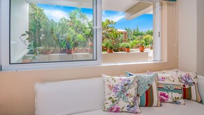 Una habitación con un gran ventanal, un sofá blanco con cojines estampados, y vista a un jardín con plantas en macetas.
