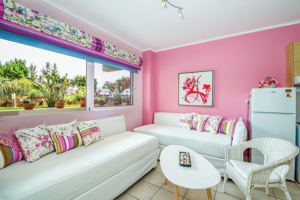 A room with two white sofas, a white table, and a wicker chair. There is a window with a view of greenery outside.