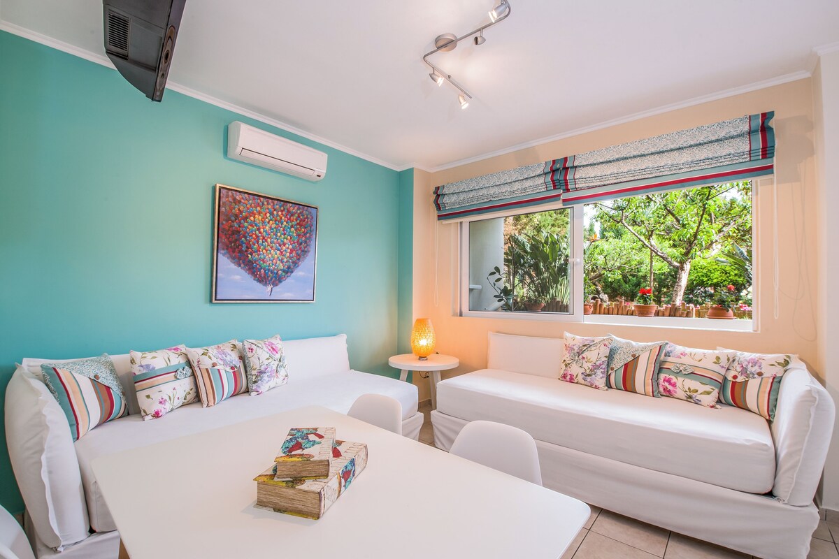 A room with two white sofas, a small table with books, a lamp, and a window with a view of greenery.