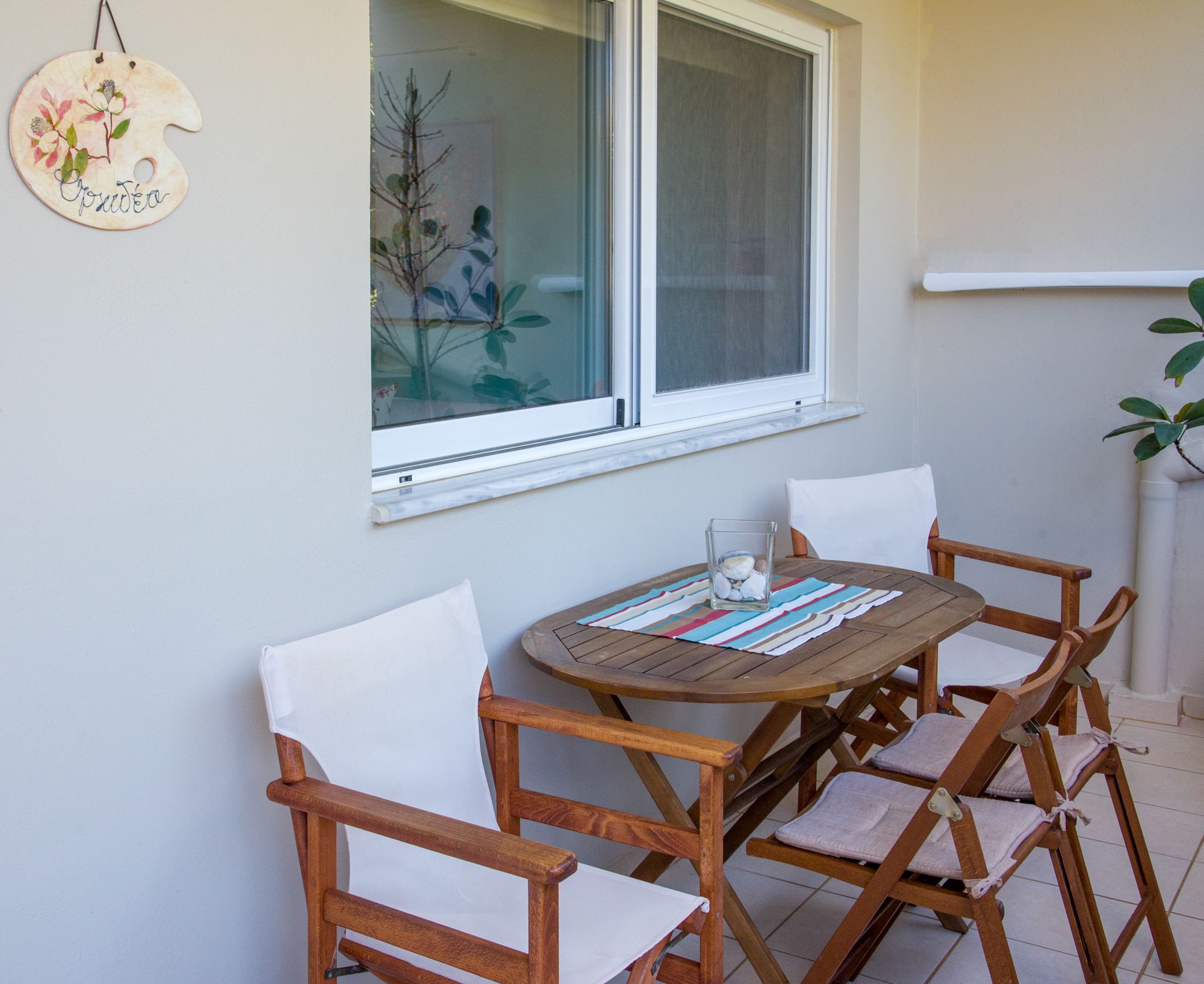 A wooden table with a glass centerpiece, surrounded by four wooden chairs with white cushions, set against a light-colored wall with a window above.