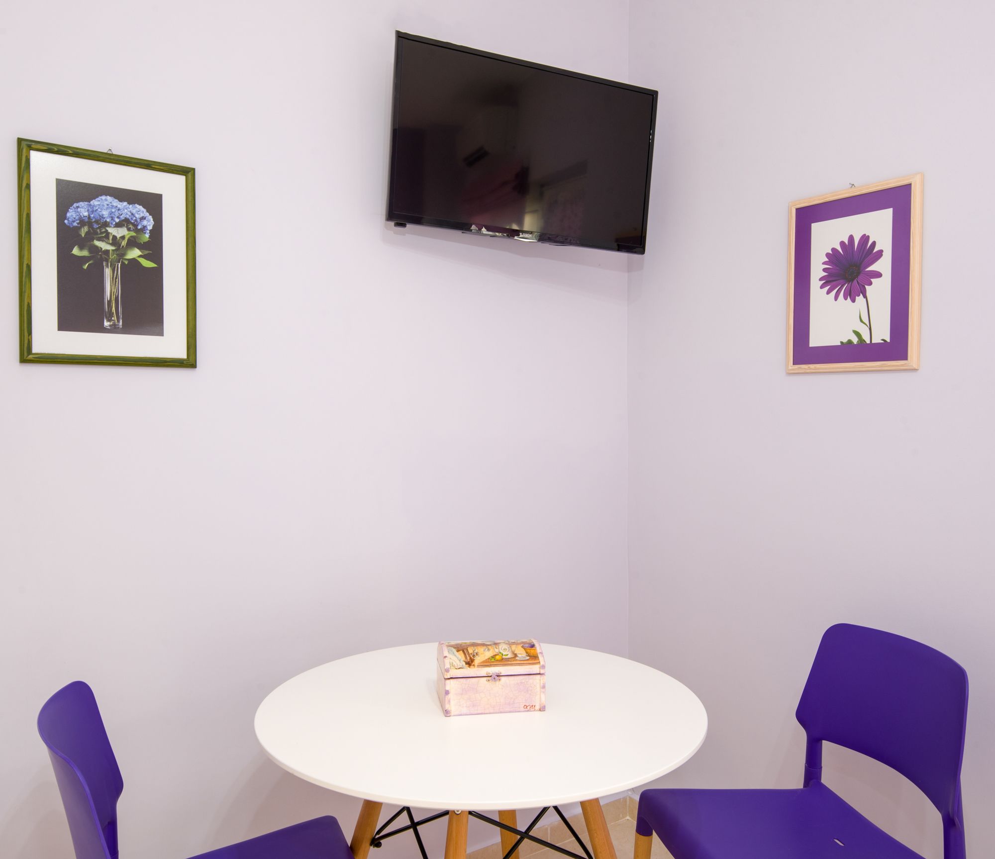 A room with a white table, two purple chairs, a television mounted on the wall, and two framed pictures on the walls.