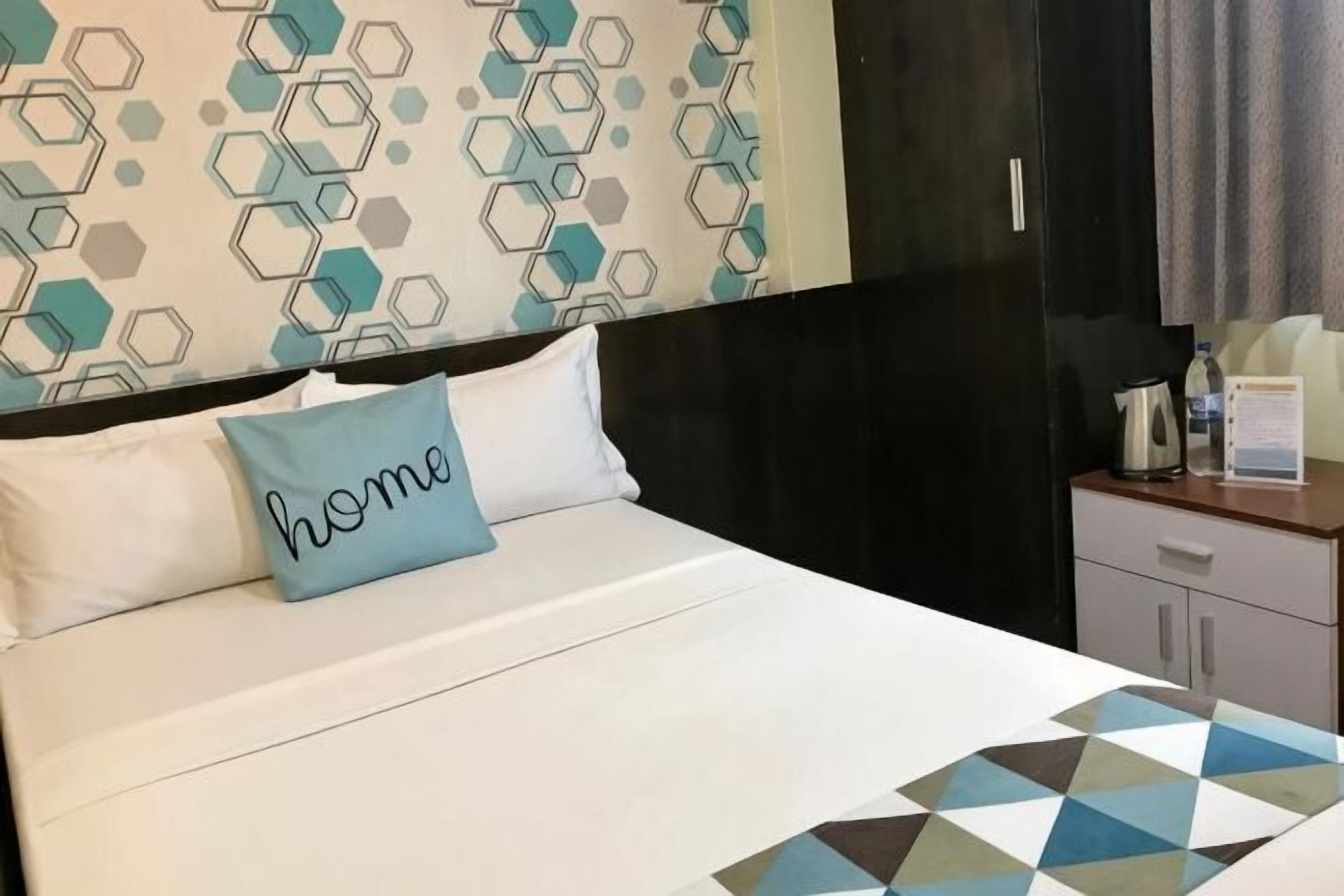 A hotel room with a bed, a pillow with 'home' written on it, a geometric-patterned bedspread, a wooden nightstand, and a black wardrobe.
