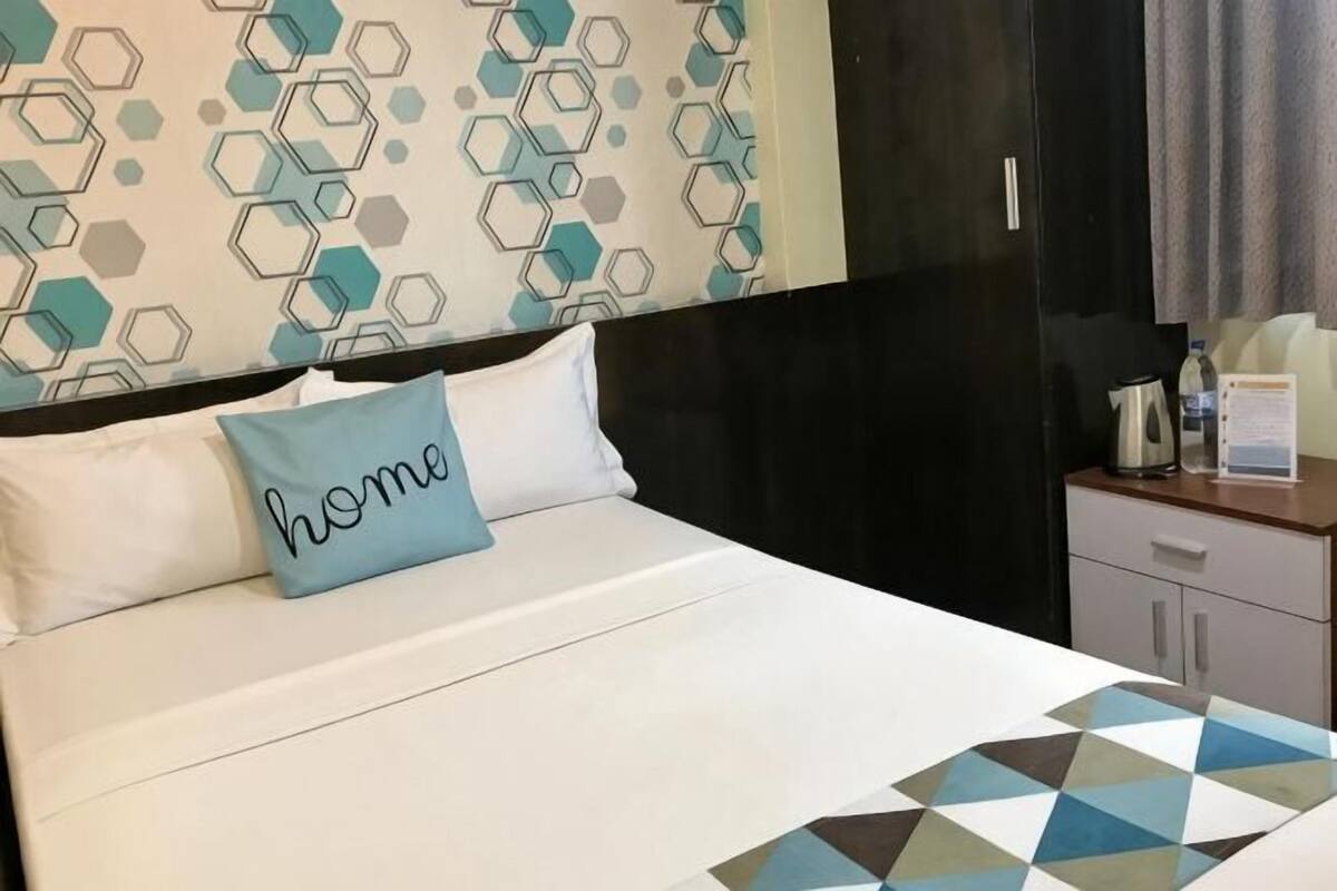 Una habitación de hotel con una cama, un almohadón con la palabra 'home' escrita, una colcha de estampado geométrico, una mesita de noche de madera y un armario negro.