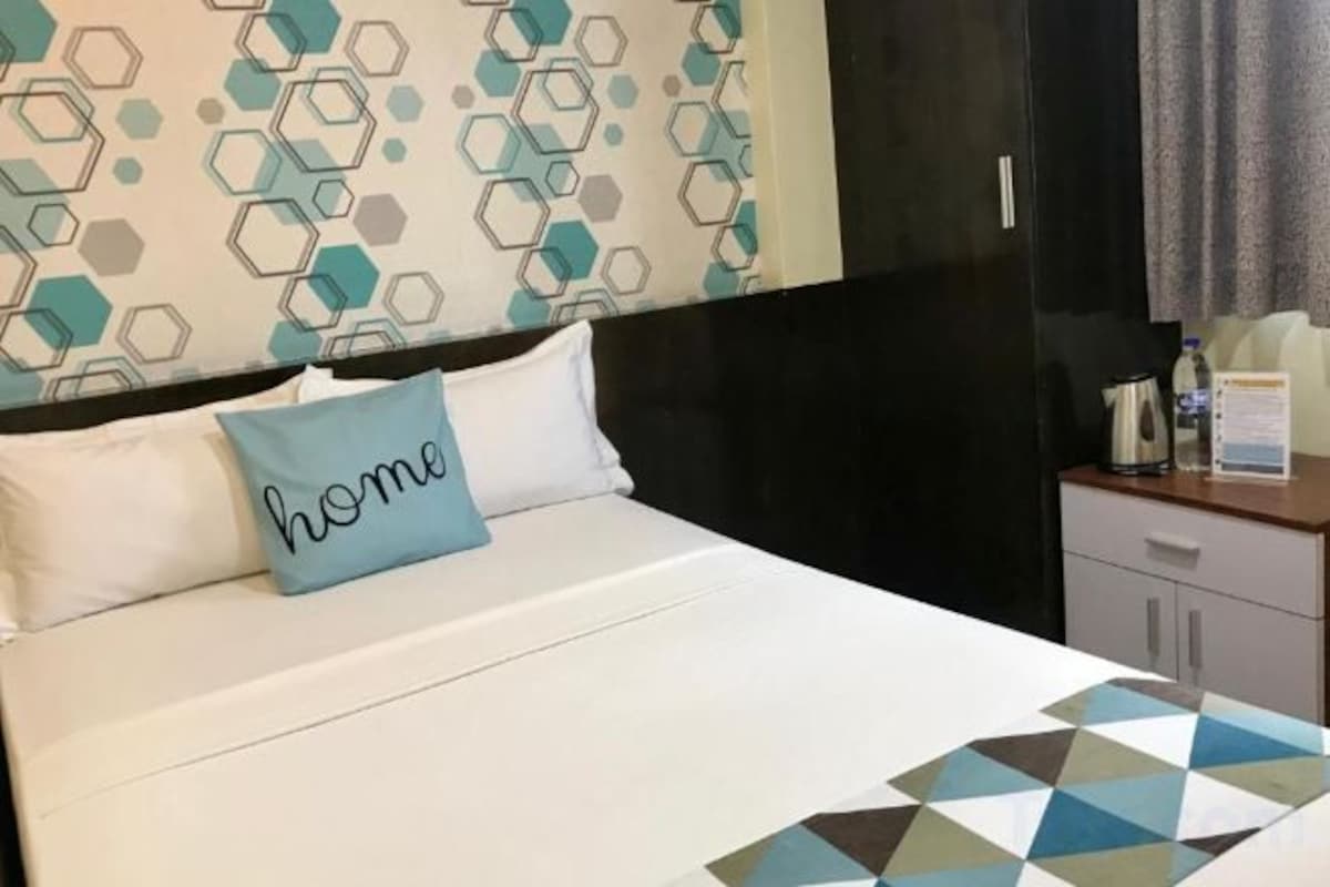 Habitación de hotel con cama, almohada con la palabra 'home', colcha de diseño geométrico, mesita de noche con tetera y cortina.