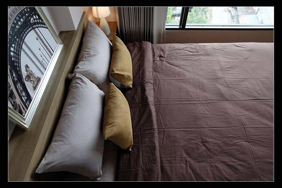 Cama bem arrumada com colcha marrom e duas almofadas, quadro na mesinha de cabeceira e janela com vista para o verde.