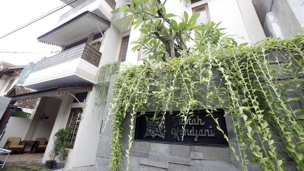 Um edifício de vários andares com varanda, uma placa com logótipo e texto, e uma árvore com trepadeiras penduradas.