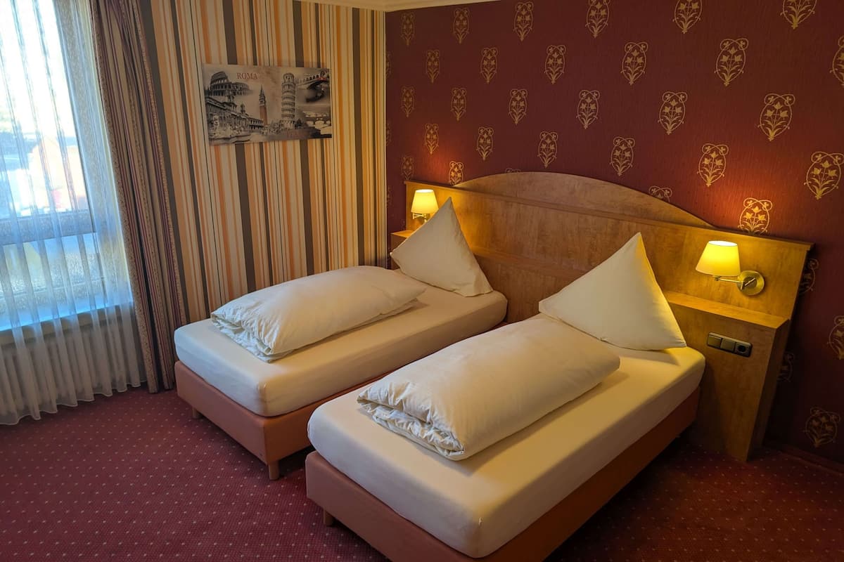 A hotel room with two single beds, a large window with curtains, a wall-mounted picture of Rome landmarks, and a patterned wallpaper.